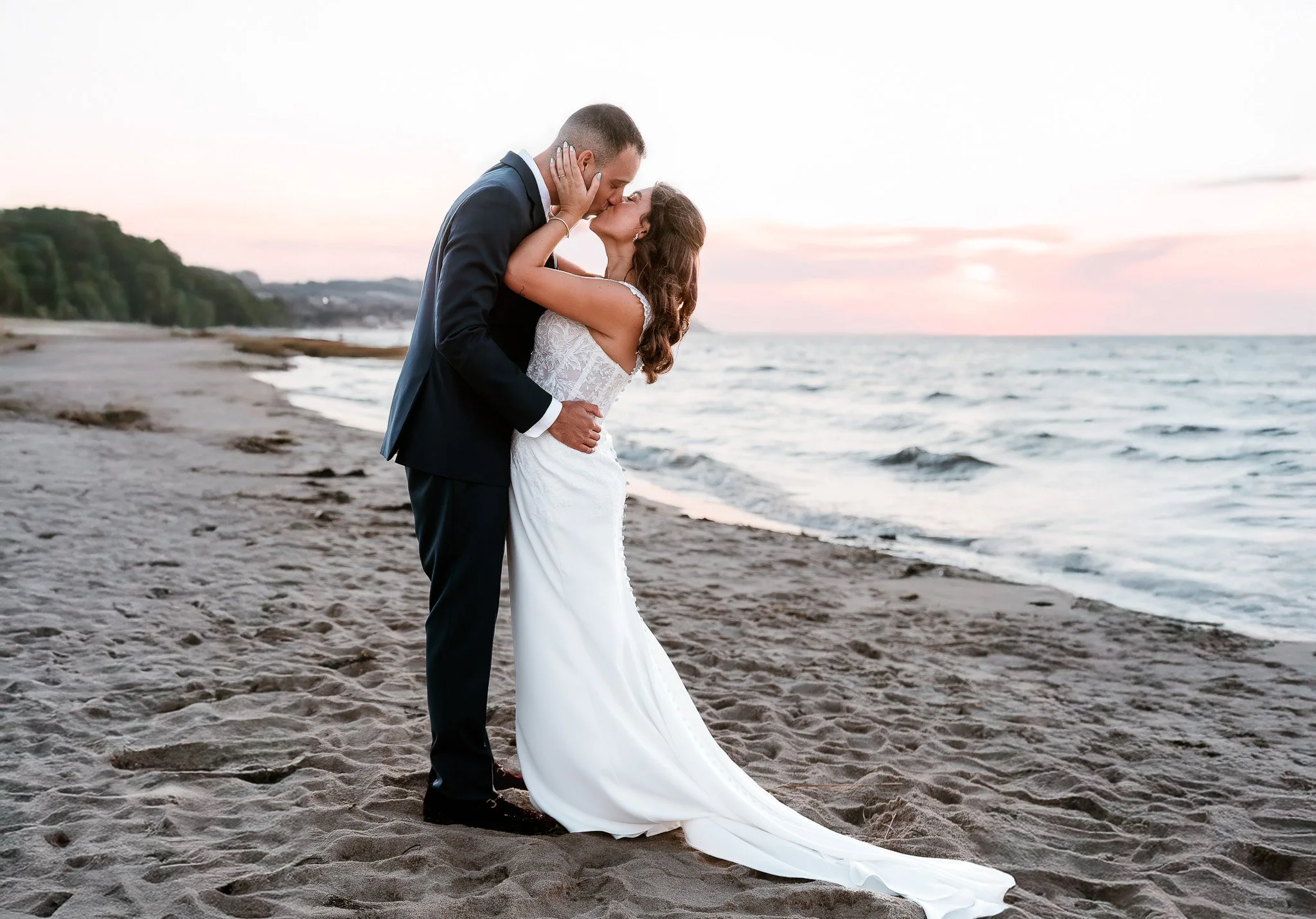 A bride and groom in wedding attire sharing a kiss on a beach at sunset, with the ocean and a distant shoreline in the background at Ocean Edge Resort