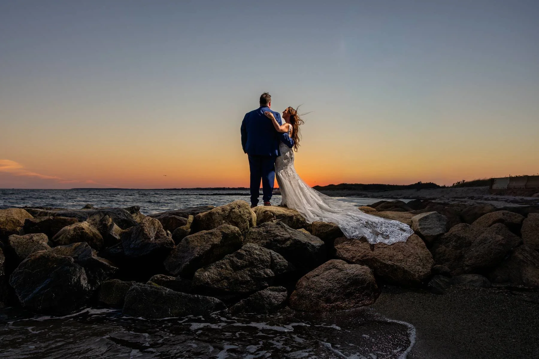 A couple in wedding attire standing on rocks by the ocean during sunset.