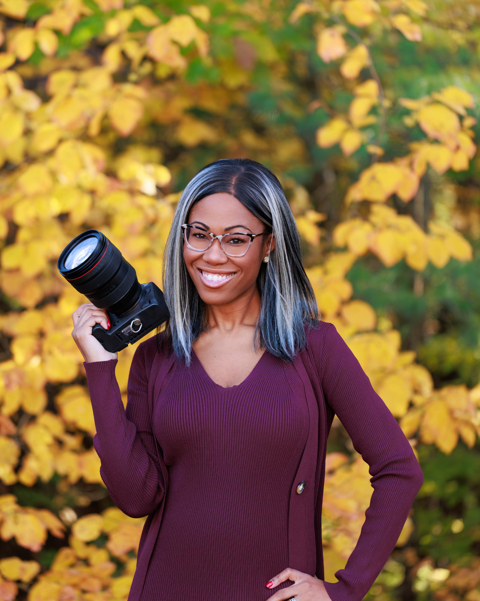 A woman with glasses and shoulder-length gray and black hair, smiling while holding a camera over her shoulder outdoors with yellow autumn leaves in the background.