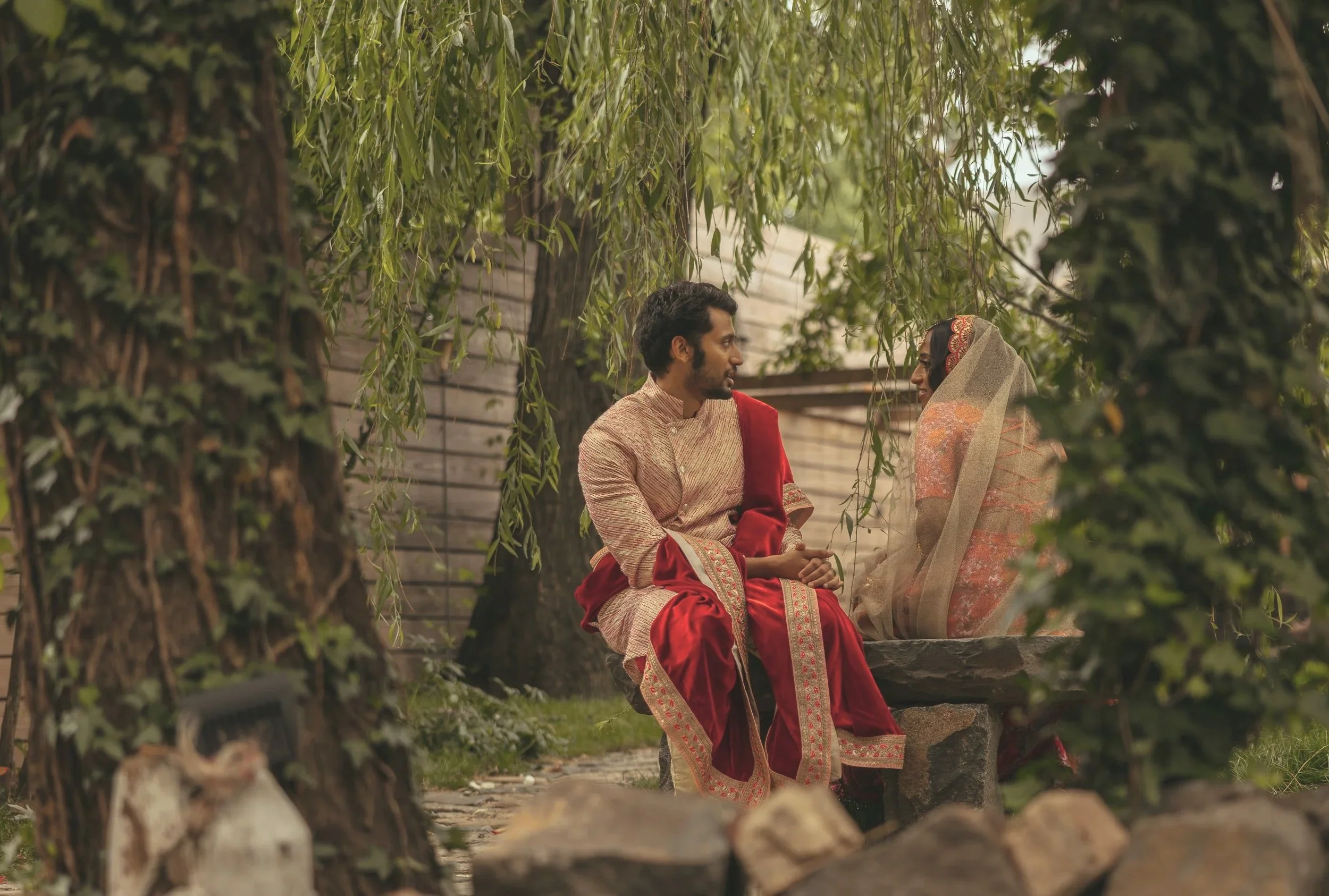A man and woman dressed in traditional Indian attire sitting on a stone bench outdoors, facing each other and smiling, with trees and a wooden fence in the background.