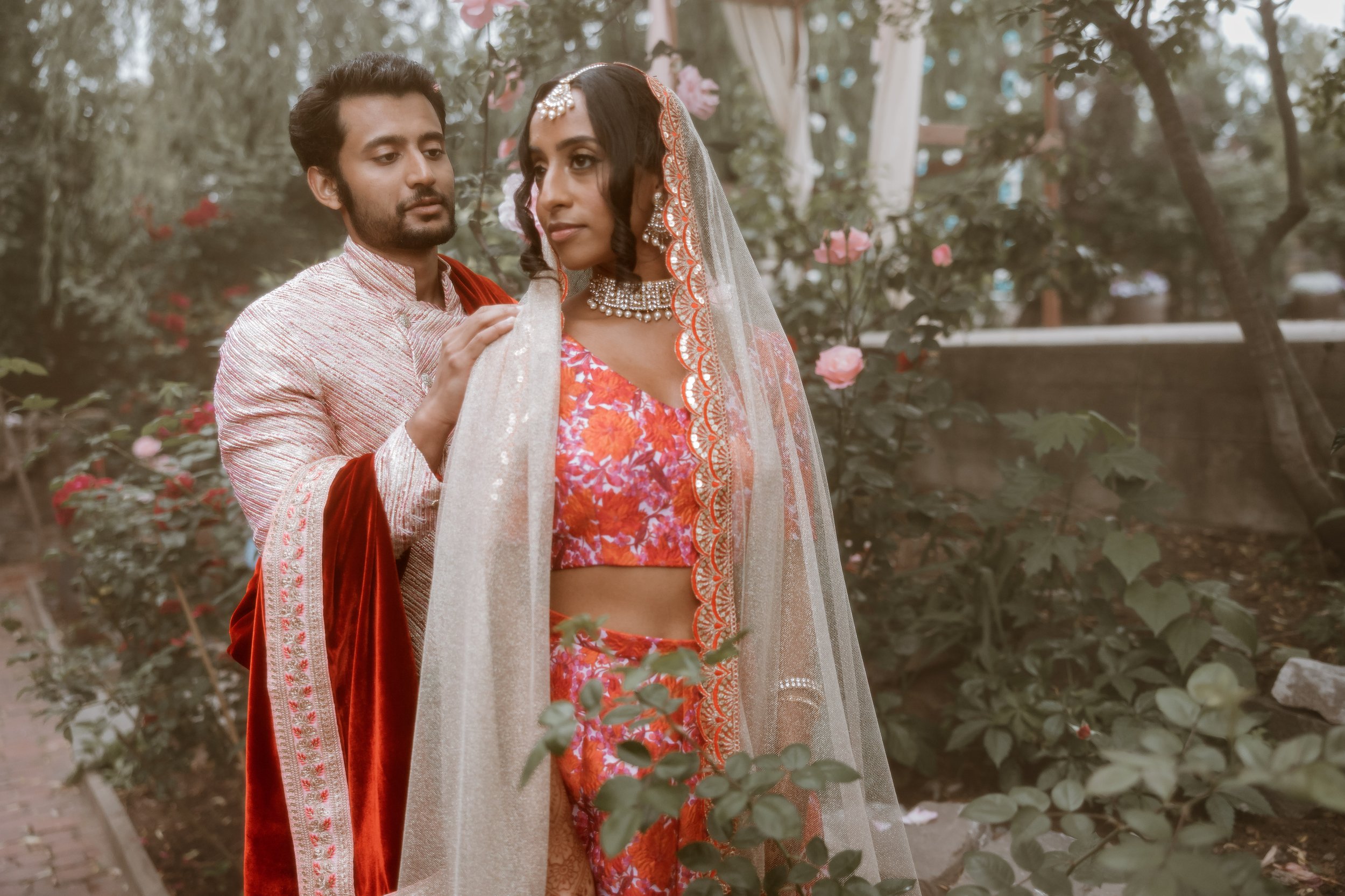 A couple dressed in traditional Indian attire, standing outdoors among rose bushes, with the man gently touching the woman's shoulder.