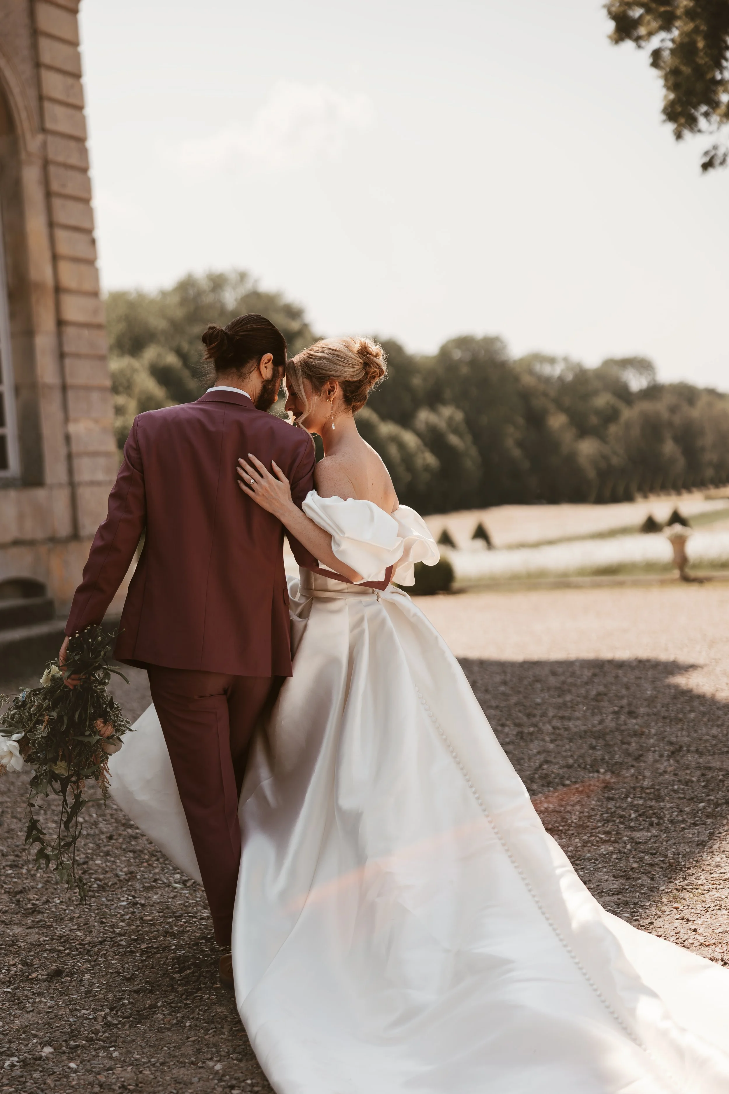 A bride and groom share a moment outside, with the bride wearing a white wedding gown with off-shoulder sleeves and the groom in a maroon suit holding a bouquet, walking close together near a stone building with greenery and trees in the background.