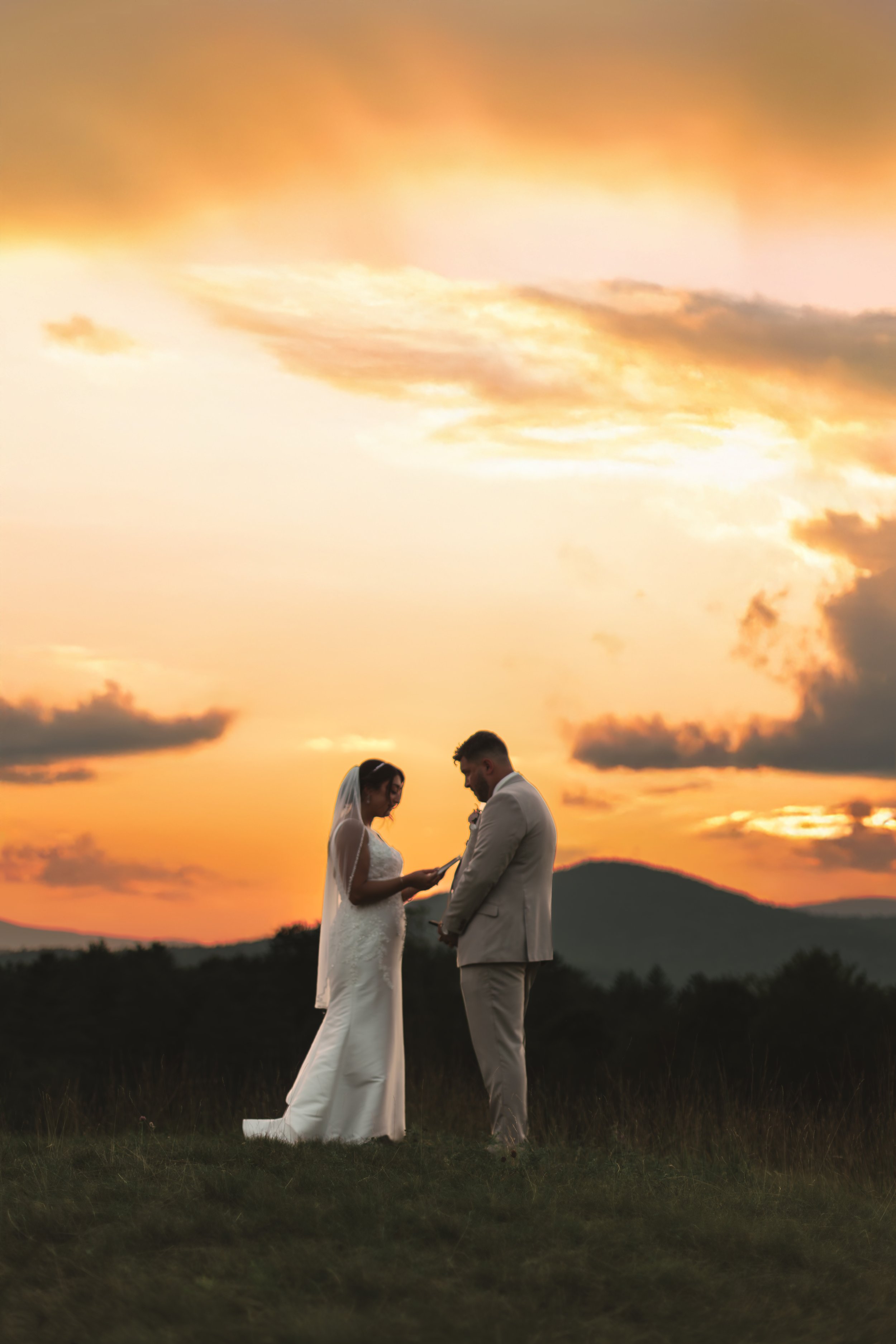 A couple dressed in wedding attire standing outdoors at sunset, with a scenic mountain backdrop.