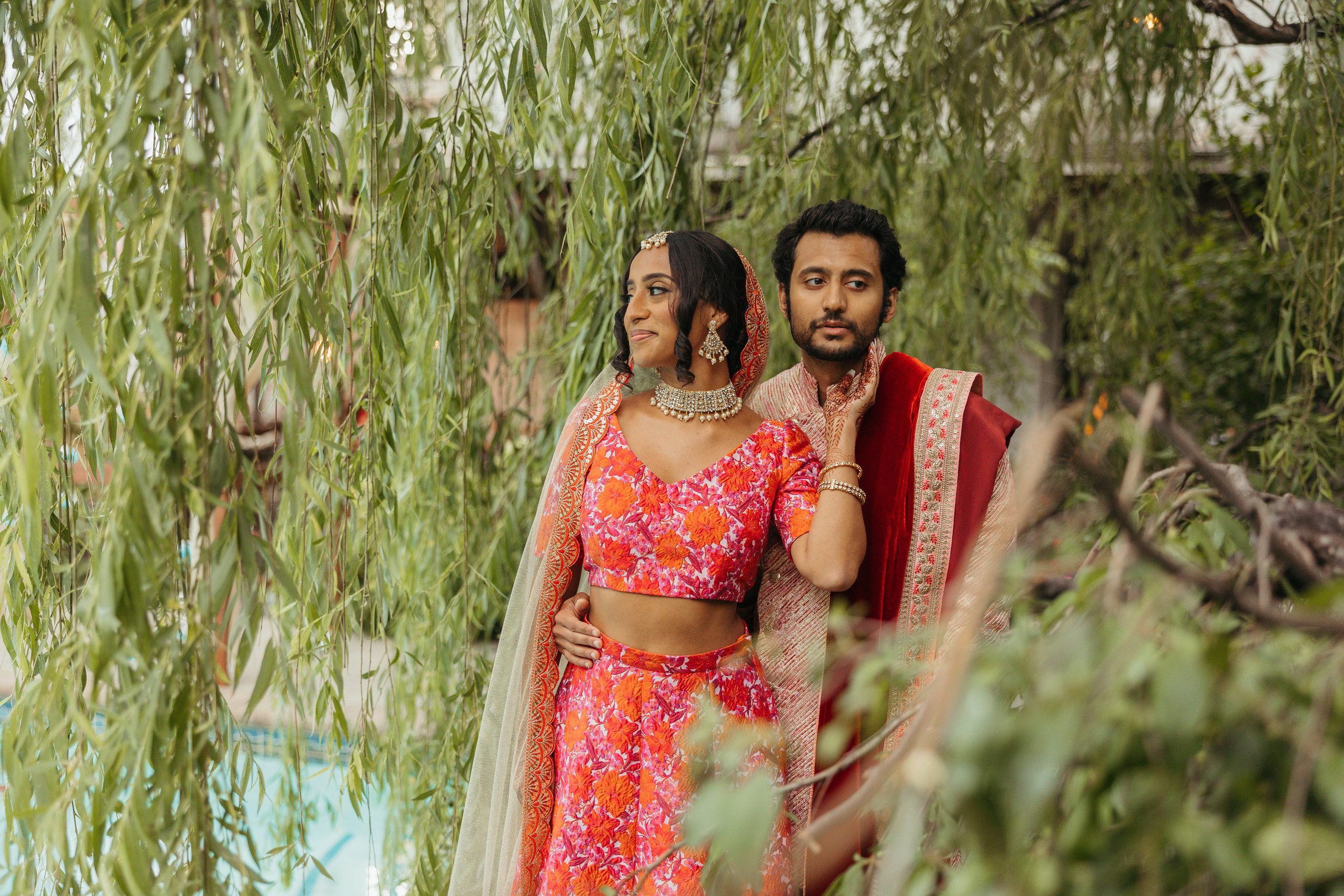 A couple dressed in traditional Indian wedding attire stands amidst green trees, with the woman smiling and looking to the side, and the man looking forward.