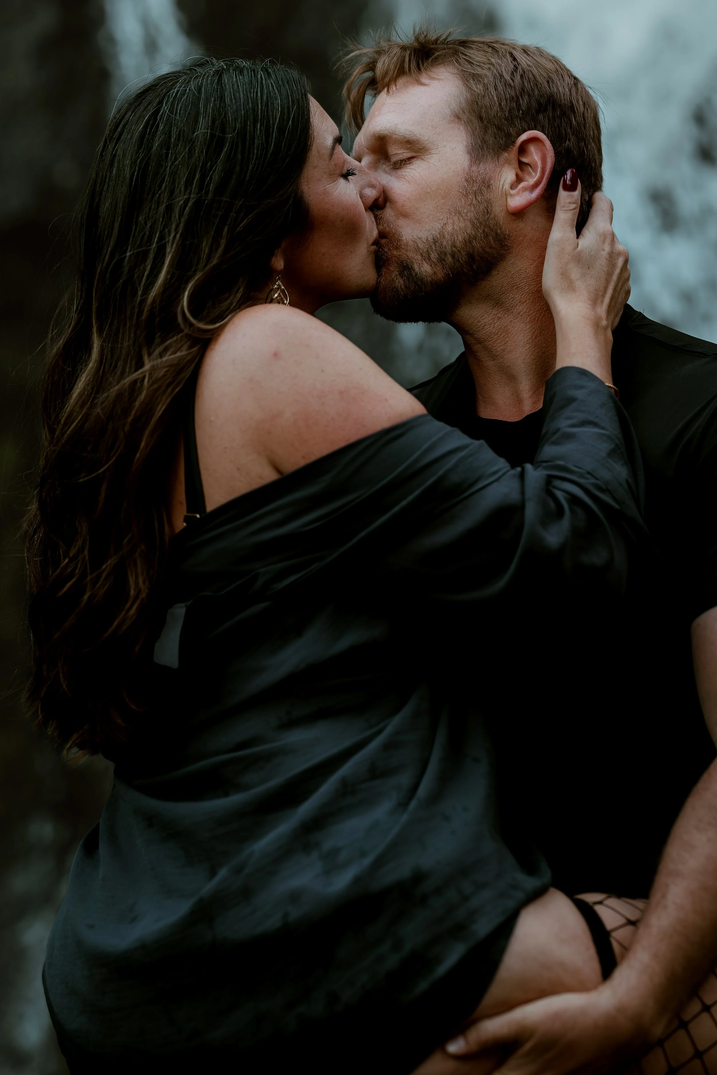 A couple sharing a passionate kiss outdoors near a waterfall.