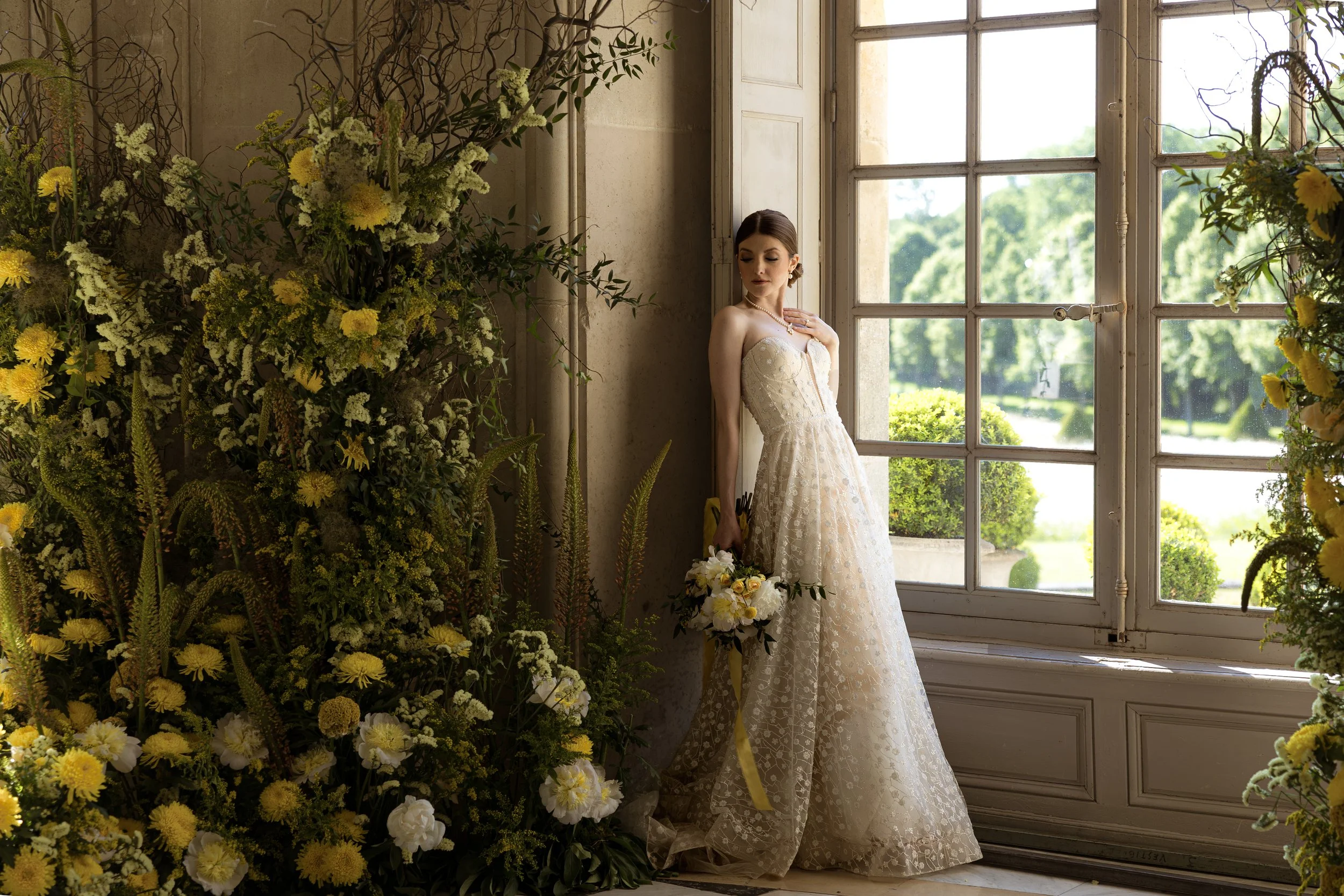 A bride in a white lace wedding dress standing indoors near a large window, surrounded by yellow and white flowers, holding a bouquet, with greenery outside visible through the window.