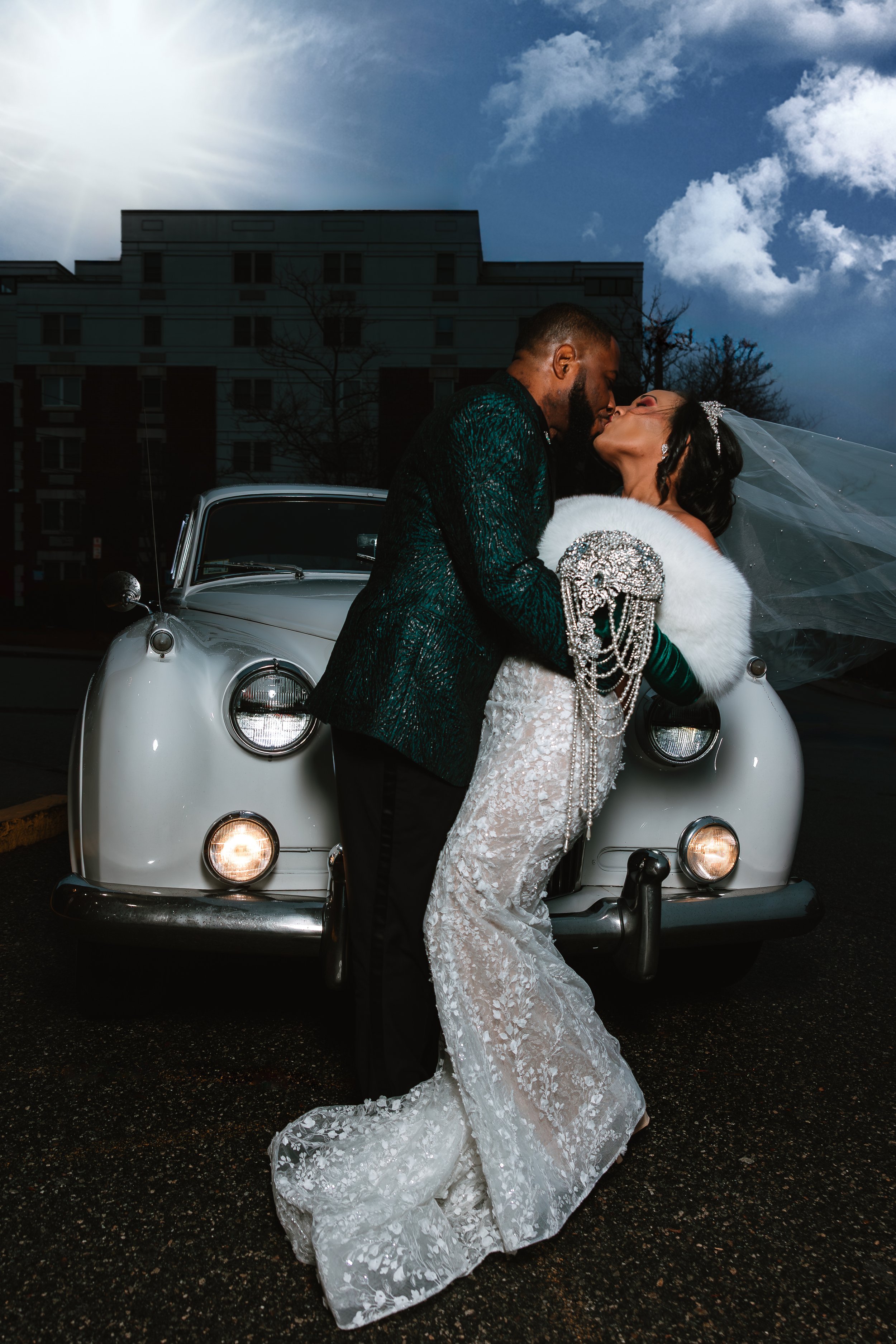 A bride and groom kissing in front of a vintage white car during their wedding.