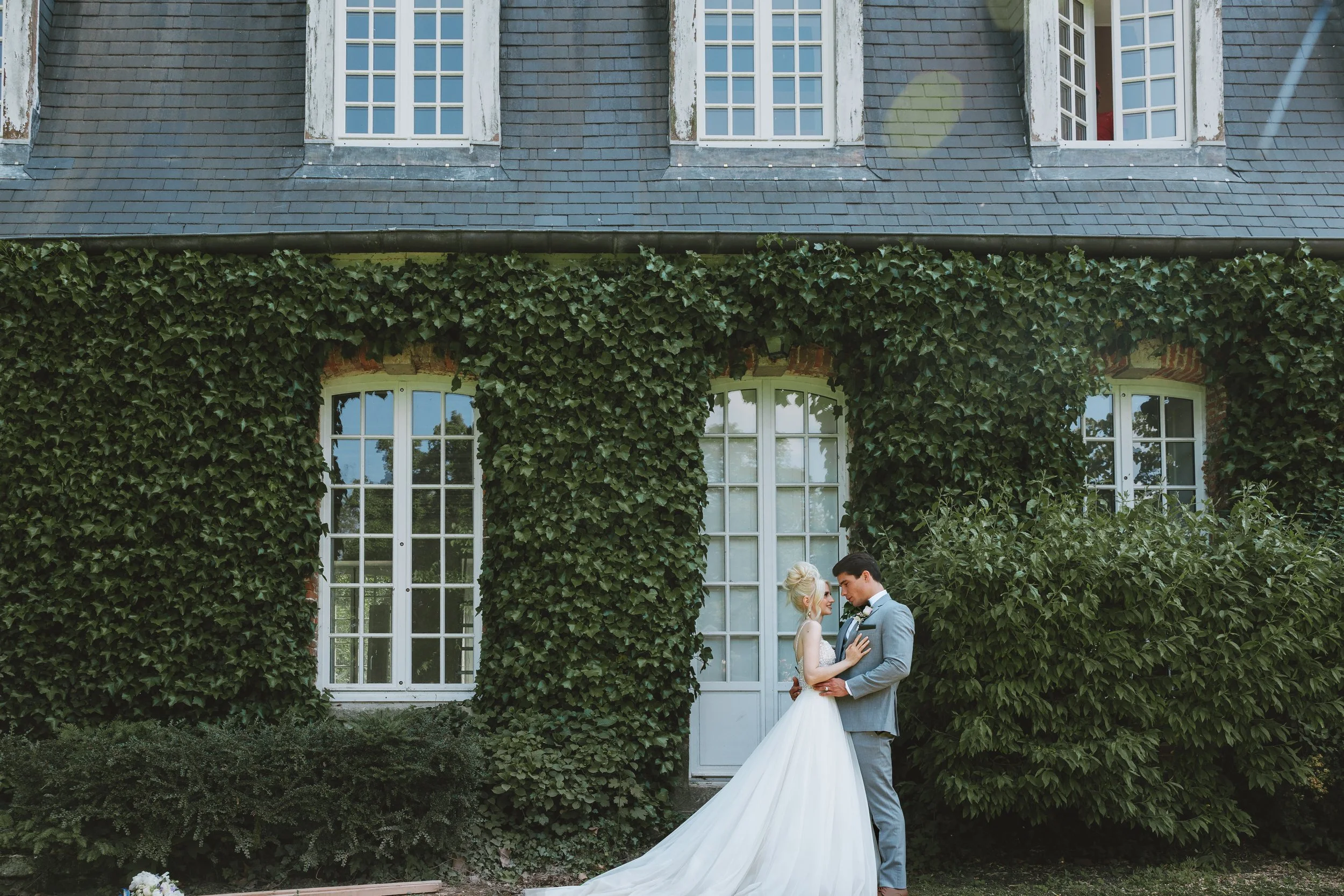 A bride and groom standing close together in front of a lush green ivy-covered building, sharing a moment.