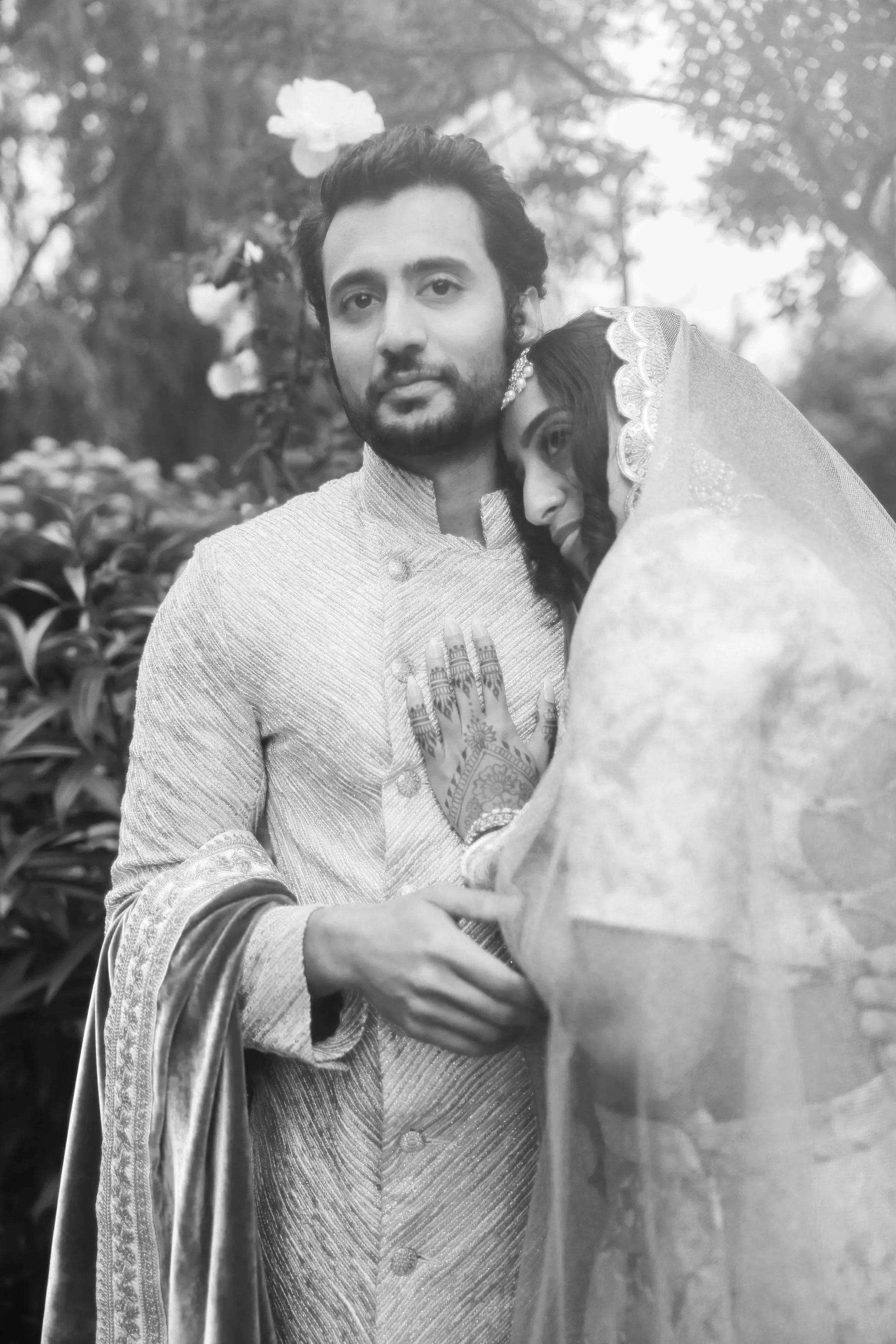 A black and white photo of a couple dressed in traditional Indian wedding attire, embracing outdoors surrounded by trees and foliage.