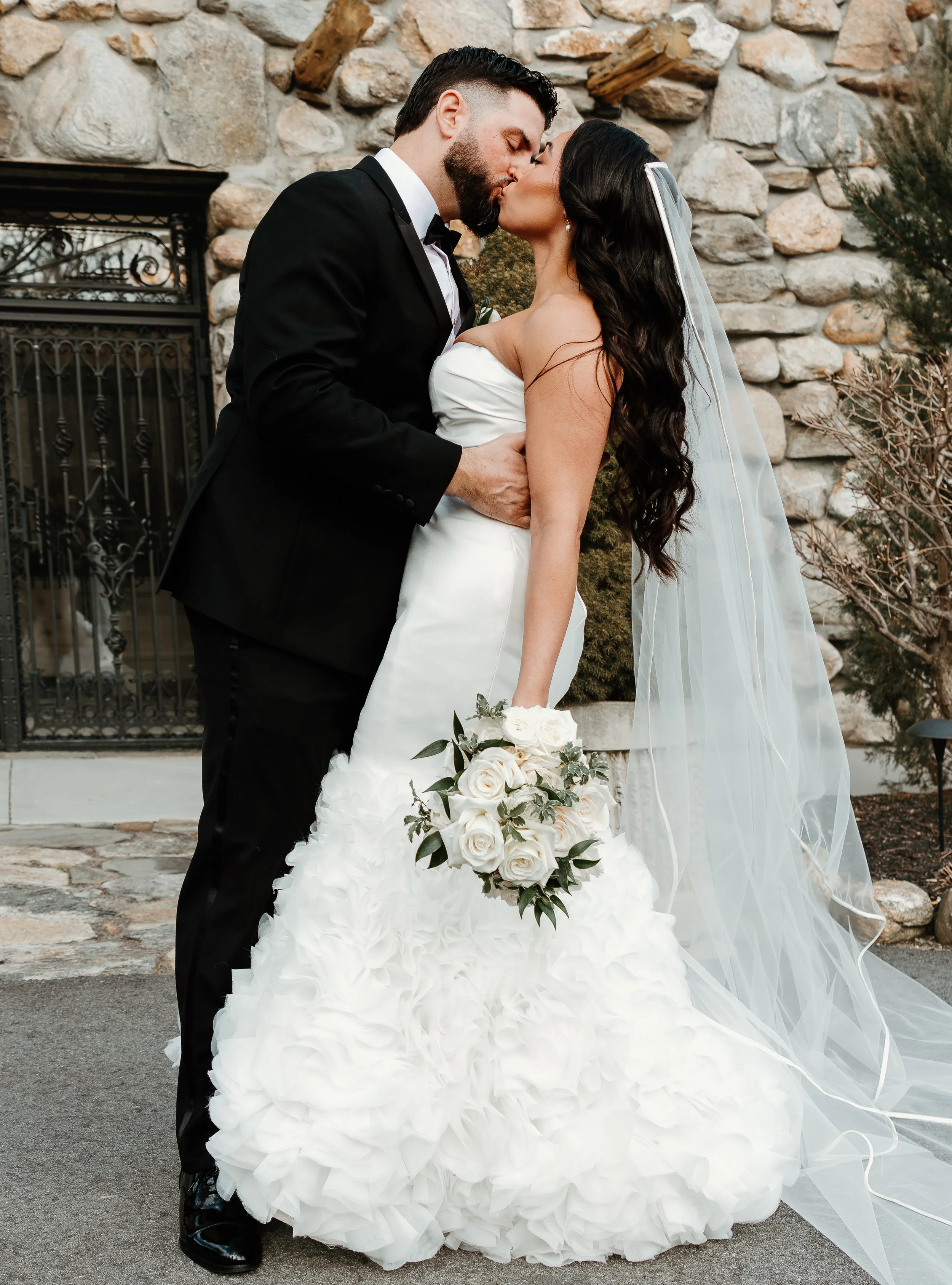 Elegant bride and groom kissing outside the stone facade at Stonehurst at Hampton Valley, featuring a strapless white wedding dress, long veil, and classic black tuxedo at a New England wedding venue.