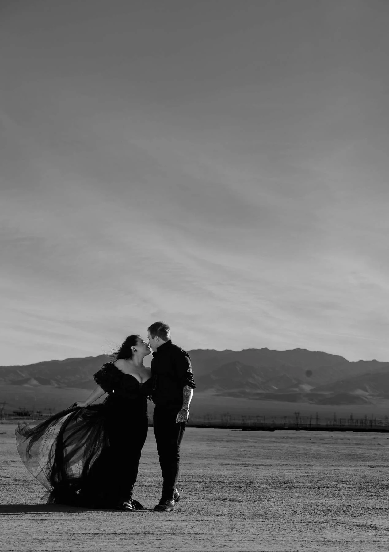 A couple kissing outdoors, woman in a flowing dress and man in dark clothing, mountain range in the background in black and white.