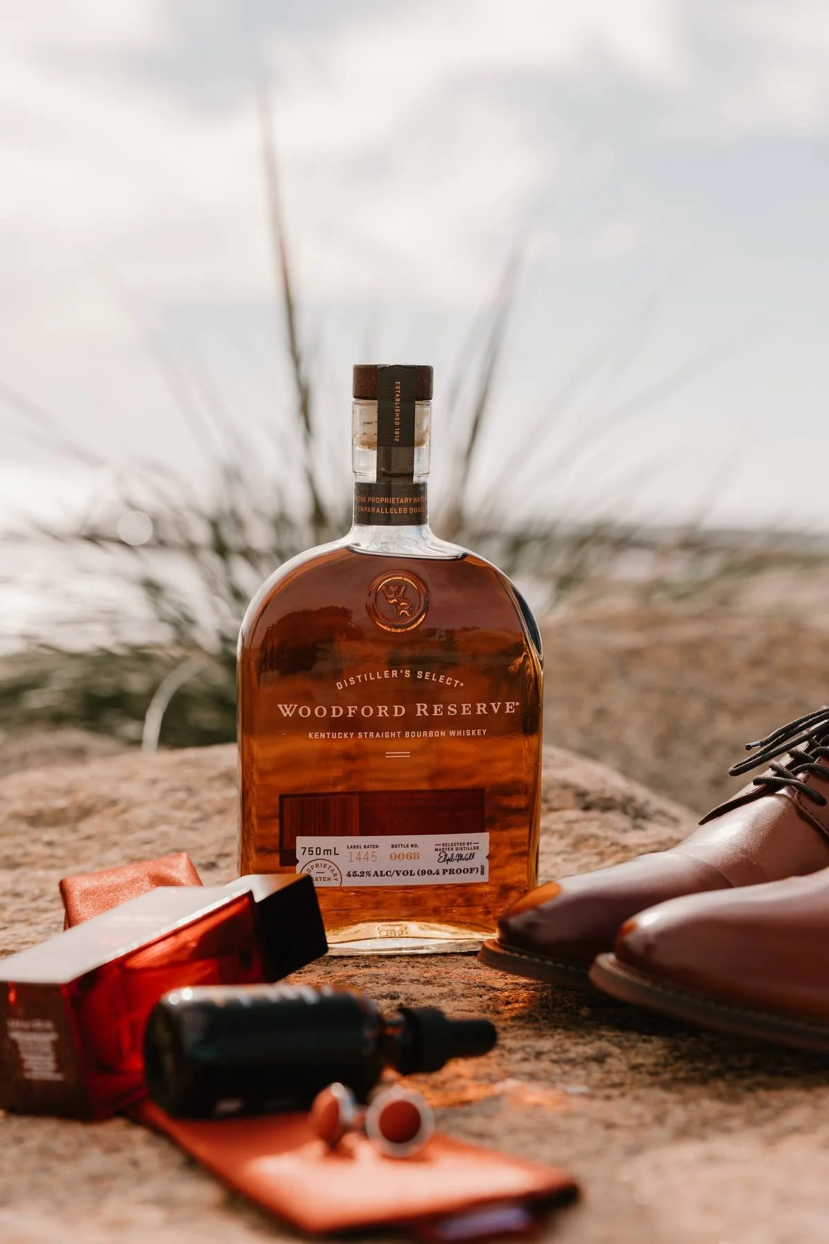 A bottle of Woodford Reserve bourbon whiskey placed on a rock surface outdoors, with a pair of pink shoes, a red box, and a small black bottle with an orange label lying nearby. The background includes blurred dry vegetation and a cloudy sky.