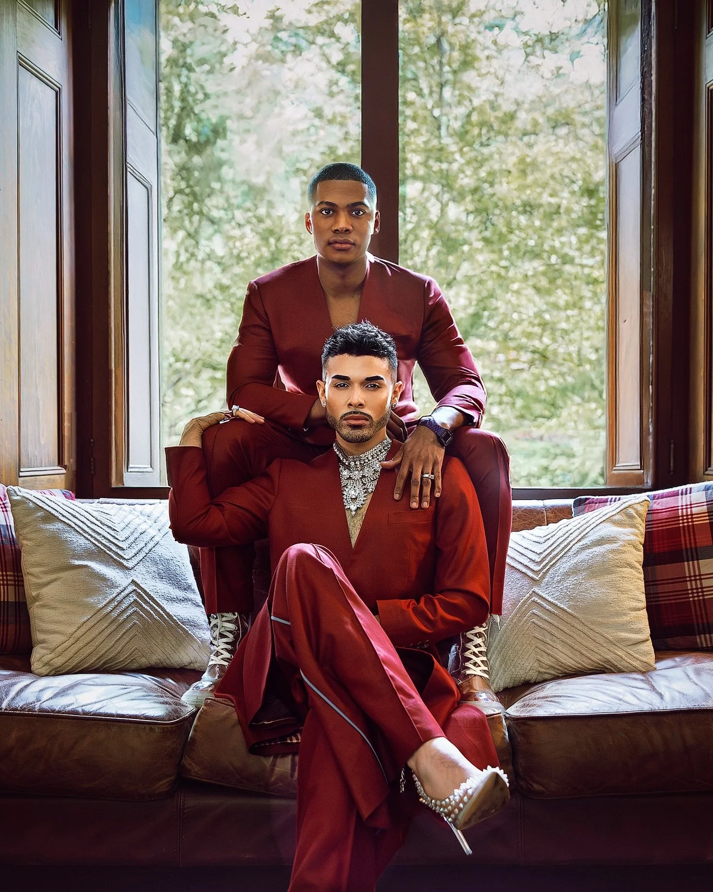 Two individuals dressed in matching maroon outfits, one sitting and the other kneeling behind on a sofa with pillows, in front of a large window with trees outside.