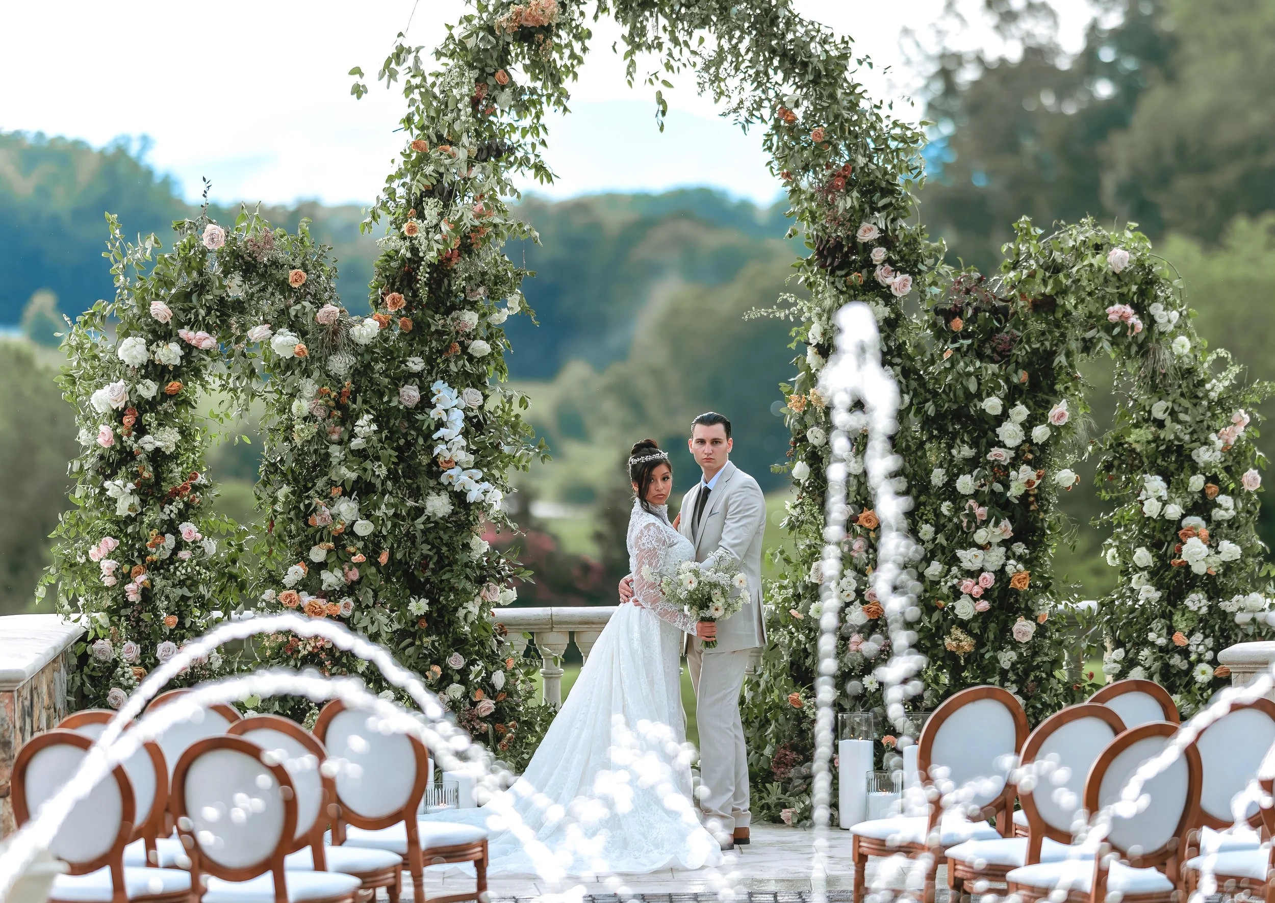 A bride and groom standing together under a floral arch during an outdoor wedding ceremony, with chairs and fountains in the foreground and a scenic landscape background.