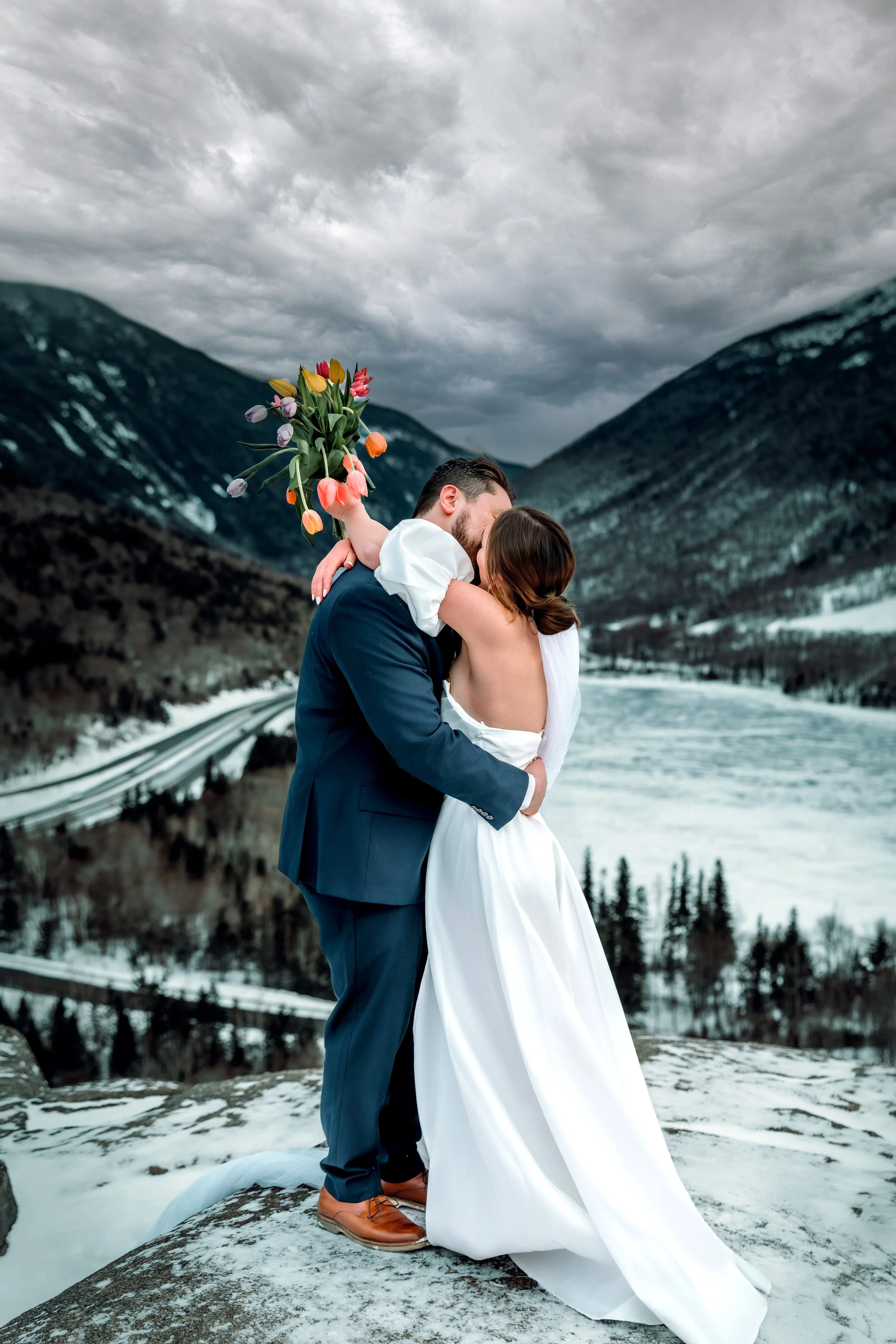 A couple in wedding attire embracing each other on a snow-covered hill with mountains and a cloudy sky in the background. The man wears a navy suit, and the woman wears a white wedding dress. The woman holds a bouquet of colorful tulips.