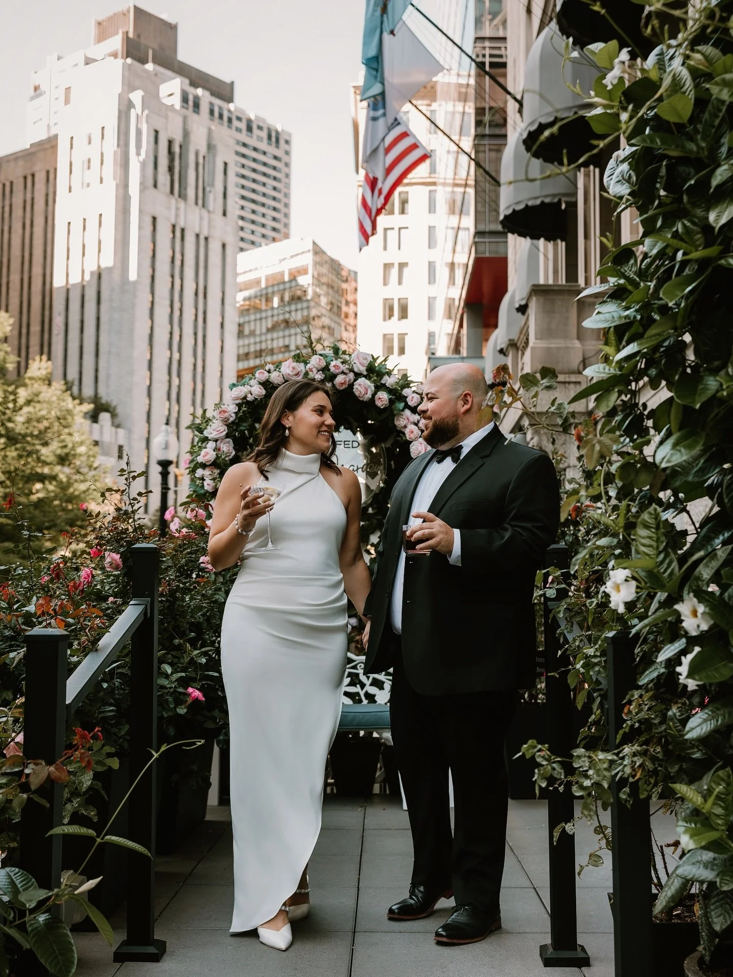 A bride and groom stand on a city rooftop garden, holding drinks and smiling at each other. The bride is in a white dress, and the groom is in a black tuxedo. There are flowers and greenery around them, with tall buildings in the background and an Am