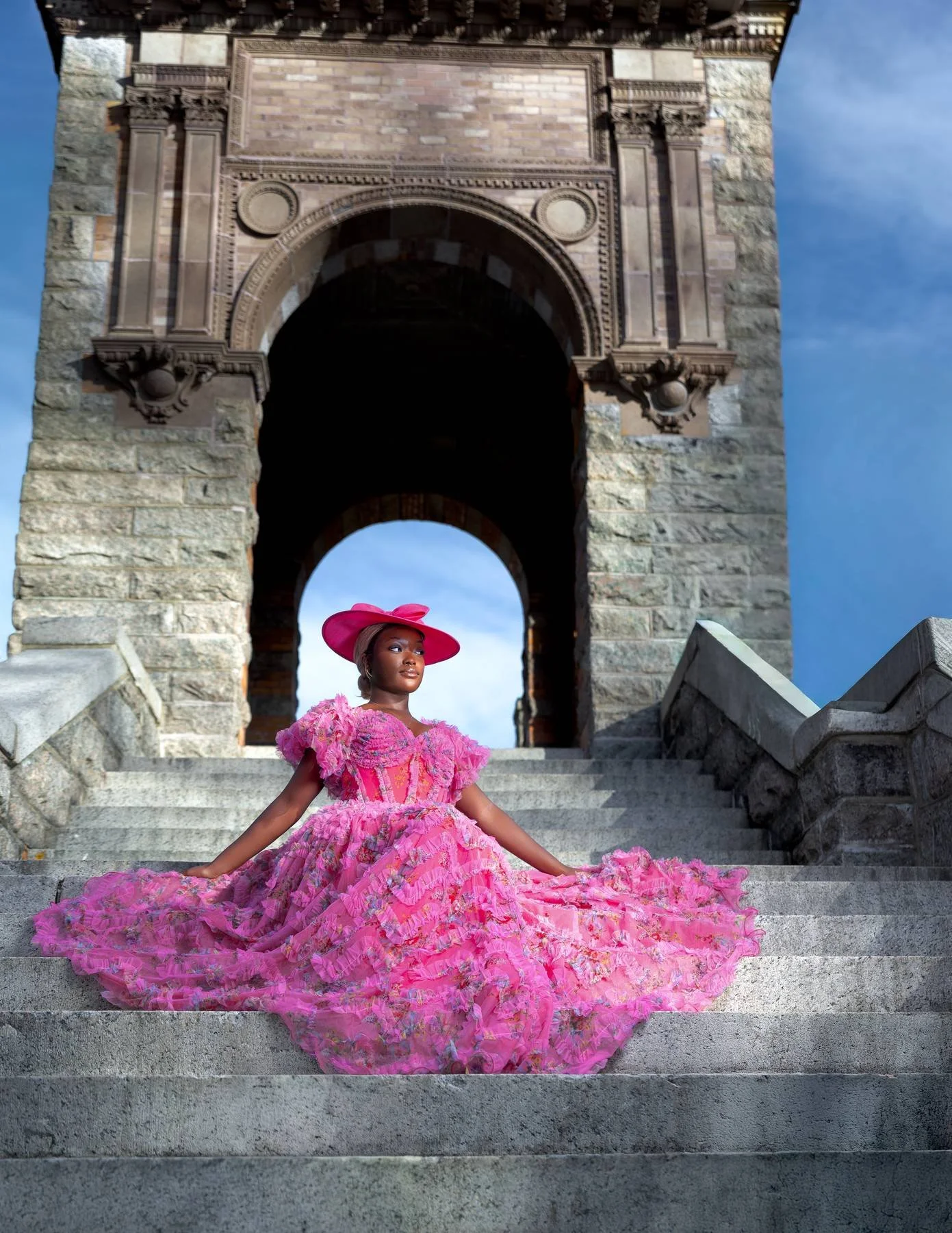 A woman in a pink, ruffled, floral gown and matching wide-brimmed hat sits on stone stairs in front of a large stone archway and historical building with sky in the background.