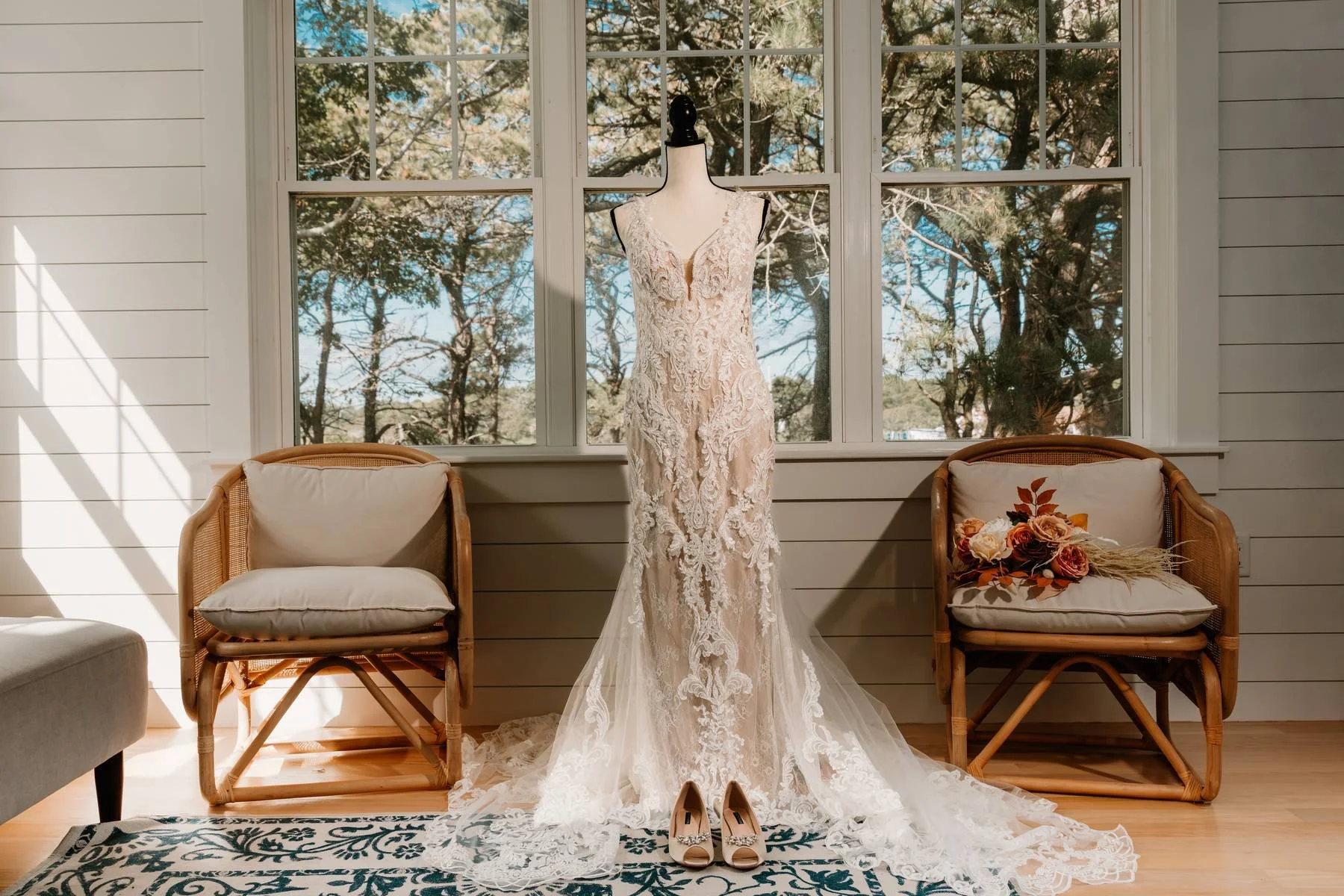 A wedding dress on a mannequin, placed on a wooden floor, with a pair of beige shoes and a bouquet of flowers in front, in front of large windows with trees outside.