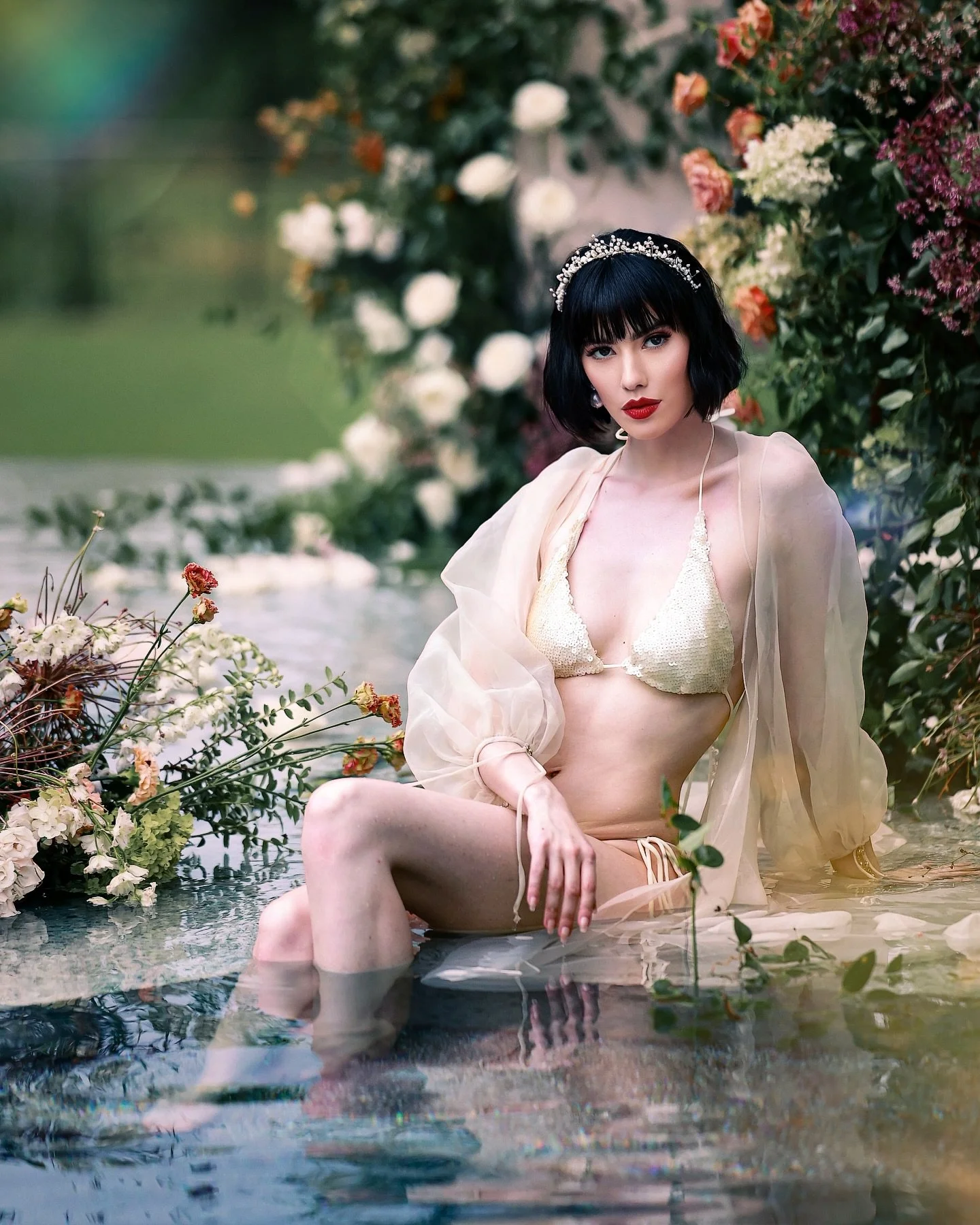 A woman with black hair and bangs wearing a pearl headband, a sheer beige cover-up, and a sequined bikini top, sitting in shallow water surrounded by flowers and greenery.