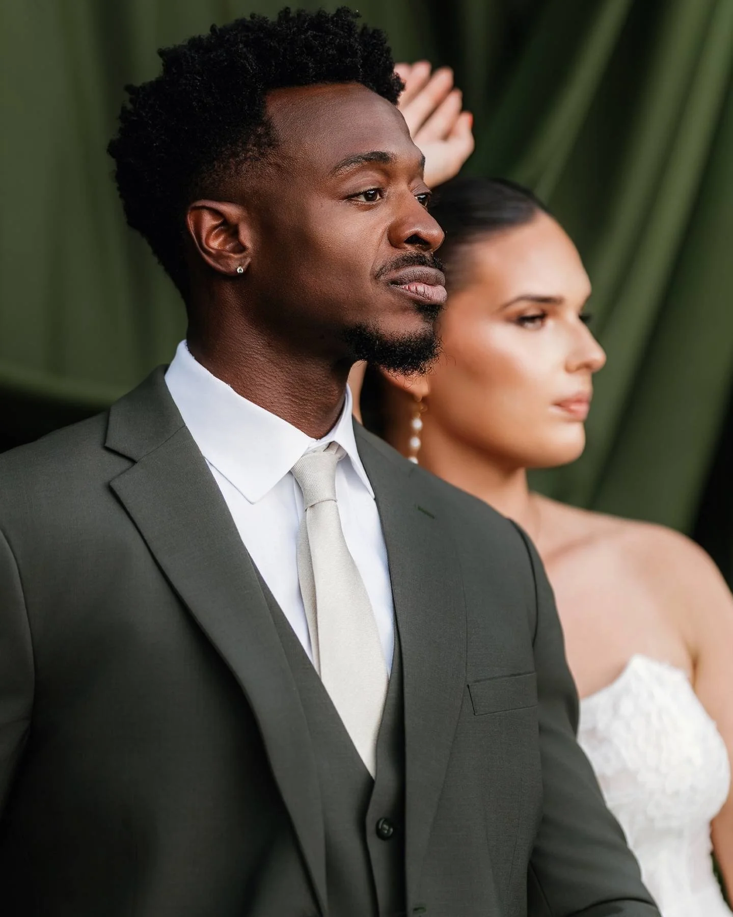 A black man in a gray suit and white tie standing with a woman in a white dress, both with serious expressions, against a green background.
