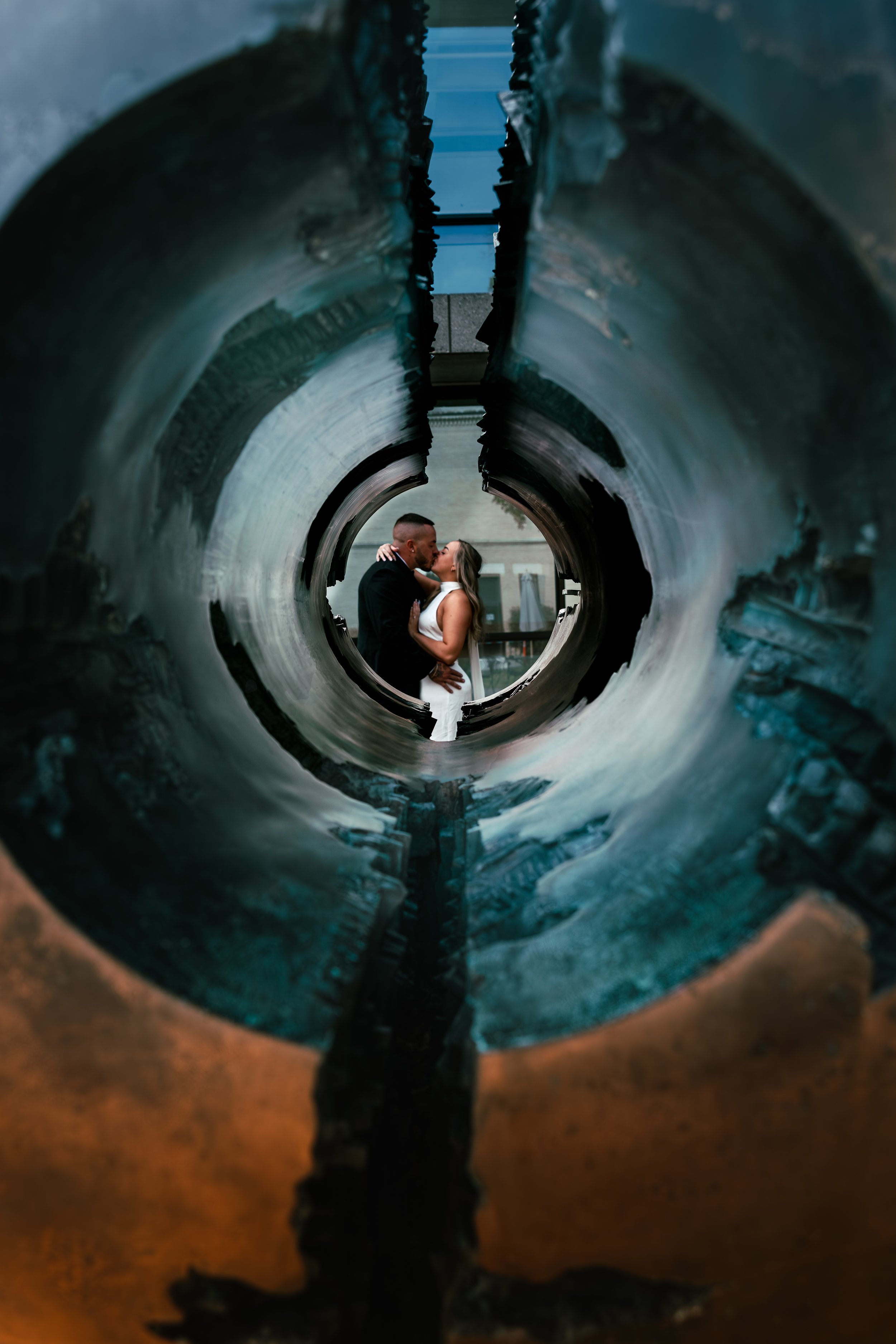 A couple kissing through a circular opening in a large metal pipe or tube, with an industrial or urban background.