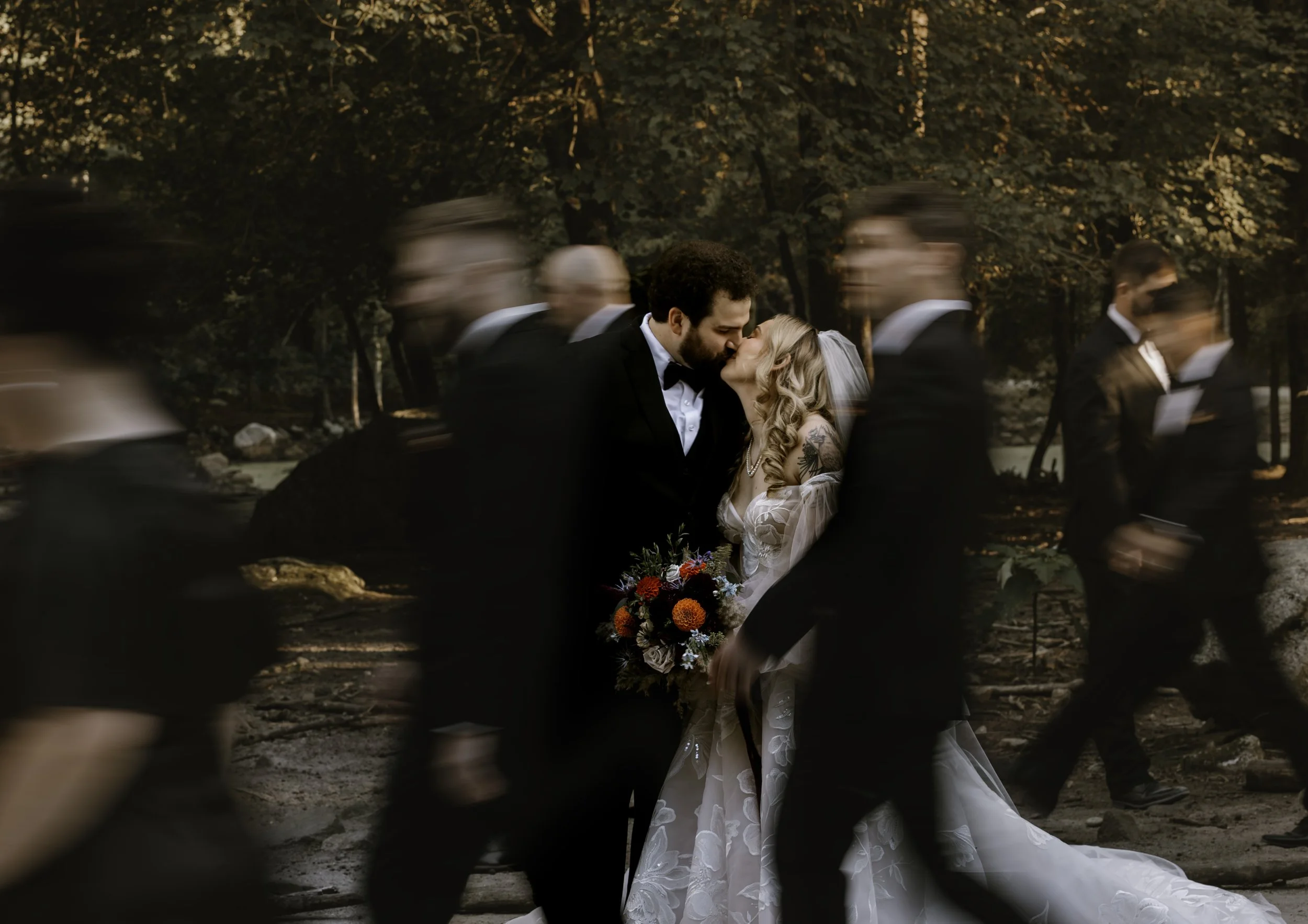 A newly married couple shares a kiss outdoors at sunset, surrounded by blurred wedding guests walking by, with a forest and river in the background.