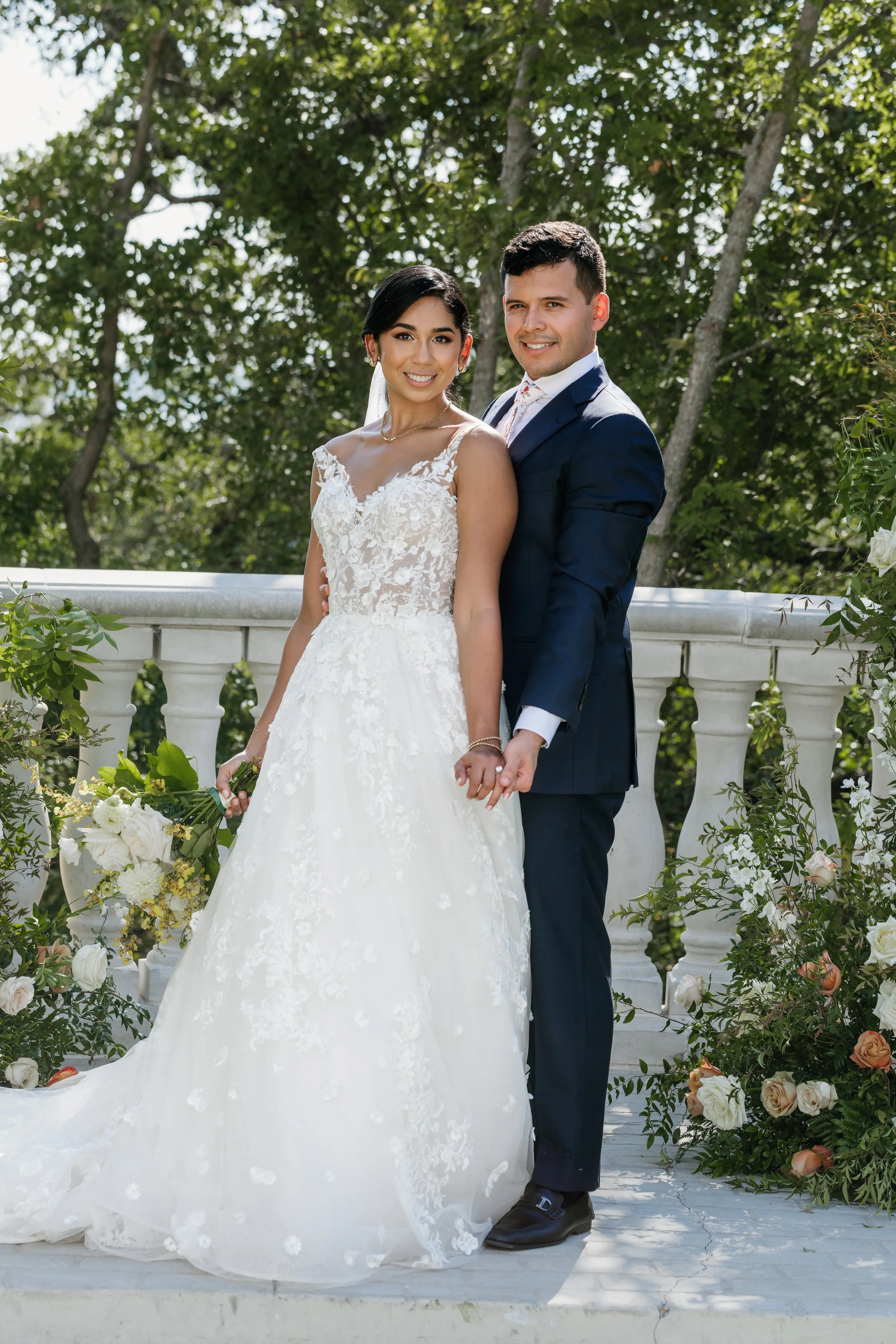 A bride and groom holding hands and smiling outdoors on their wedding day, with greenery and flowers in the background.