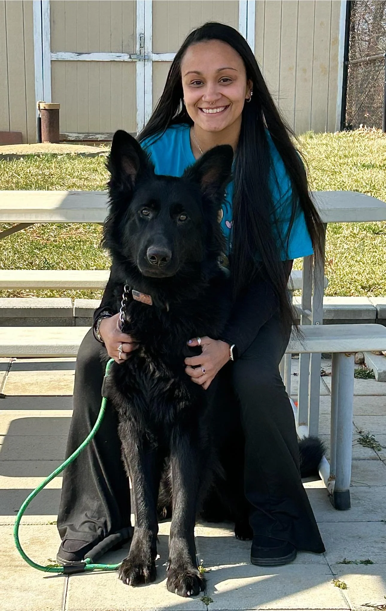 A woman with long dark hair smiling and sitting on a bench while holding a black German Shepherd dog outdoors in a yard.