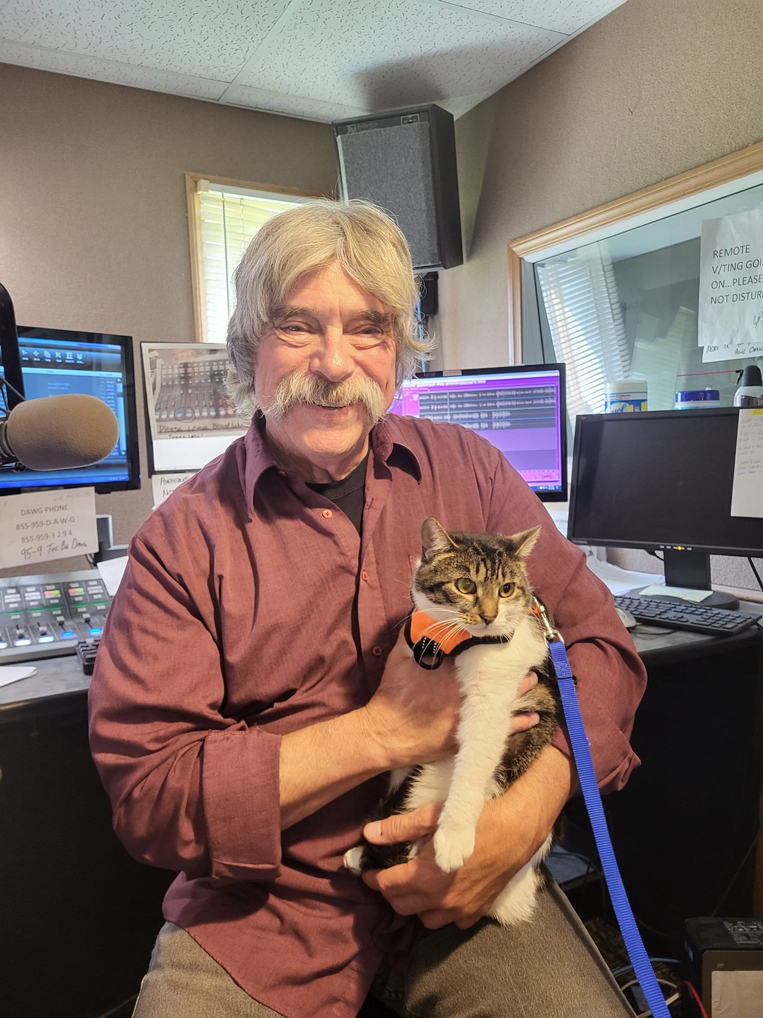 A man with gray hair and a mustache sitting in a radio station, holding a tabby cat. The man is smiling, and the cat is looking to the side. There are multiple computer monitors and audio equipment in the background.