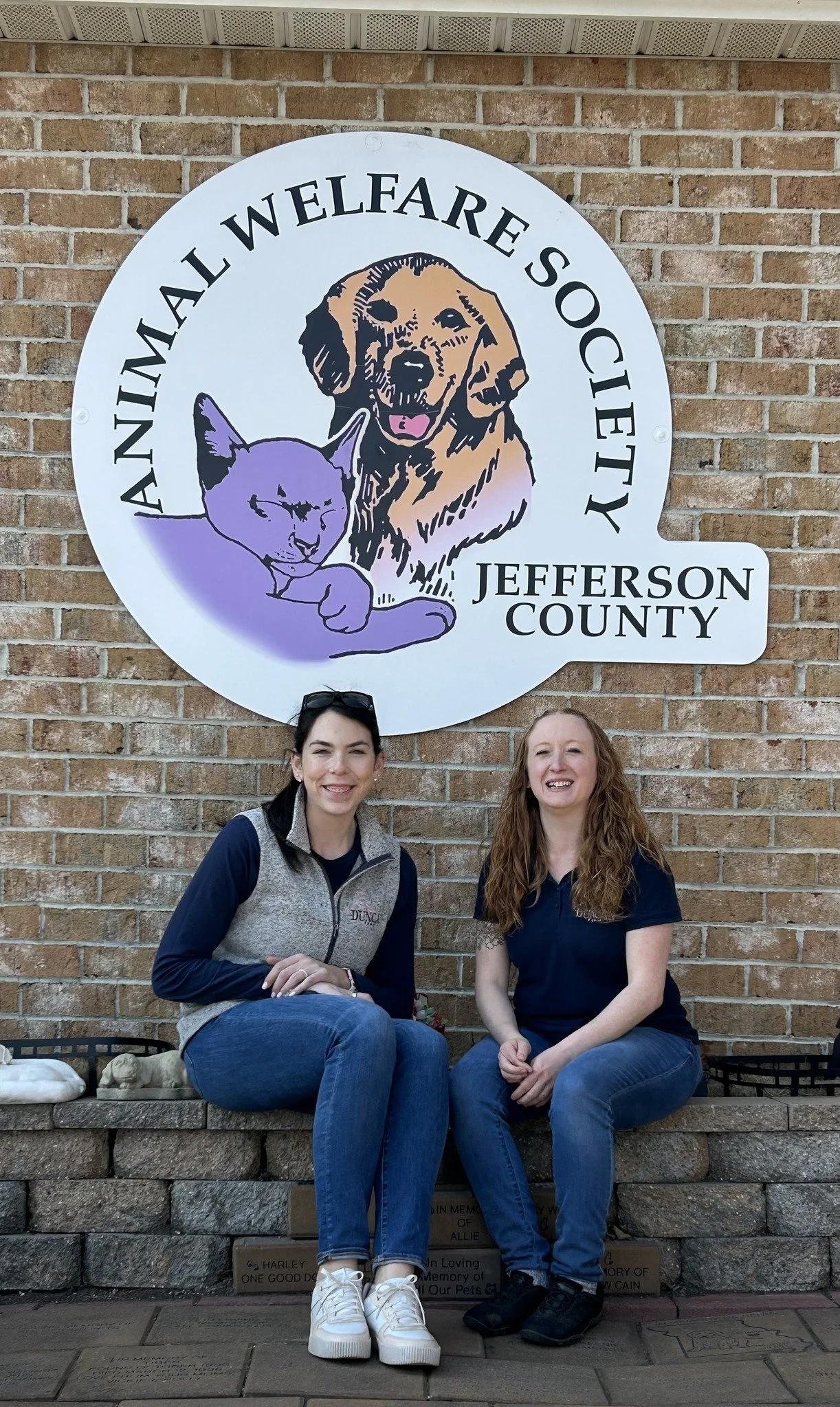 Two women sitting on a brick ledge in front of a sign for the Animal Welfare Society of Jefferson County, featuring a dog and a cat illustration.