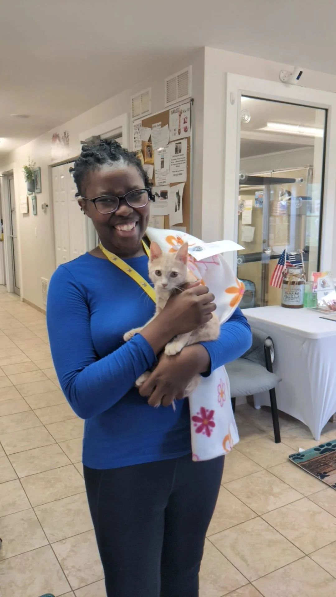 A woman smiling and holding an orange tabby kitten indoors. She is wearing glasses, a blue long-sleeve shirt, and a yellow lanyard. The background includes a bulletin board, a table with American flags, and glass doors.