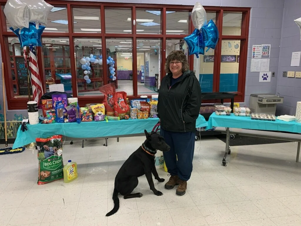 A woman and a black dog sitting in front of a table with pet supplies at an indoor event, decorated with blue and white star-shaped balloons.