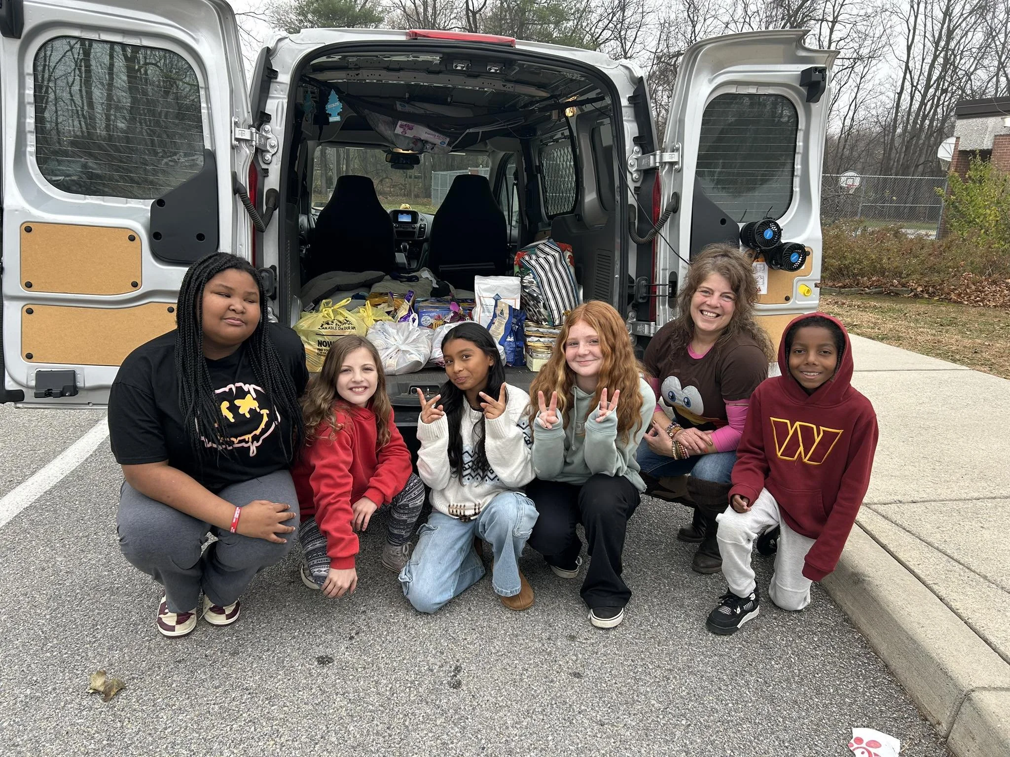Group of six children and one adult posing in front of a van filled with donations, outside on a cloudy day.