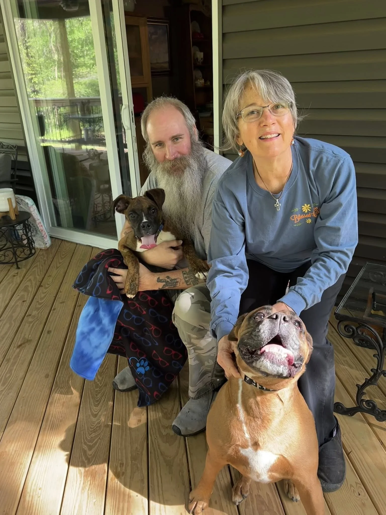 A man with a long beard, a woman with glasses, and two dogs on a wooden porch. The woman is holding a happy, smiling dog and the man is holding a small dog. The scene is bright and cheerful, with greenery visible through a sliding glass door behind t