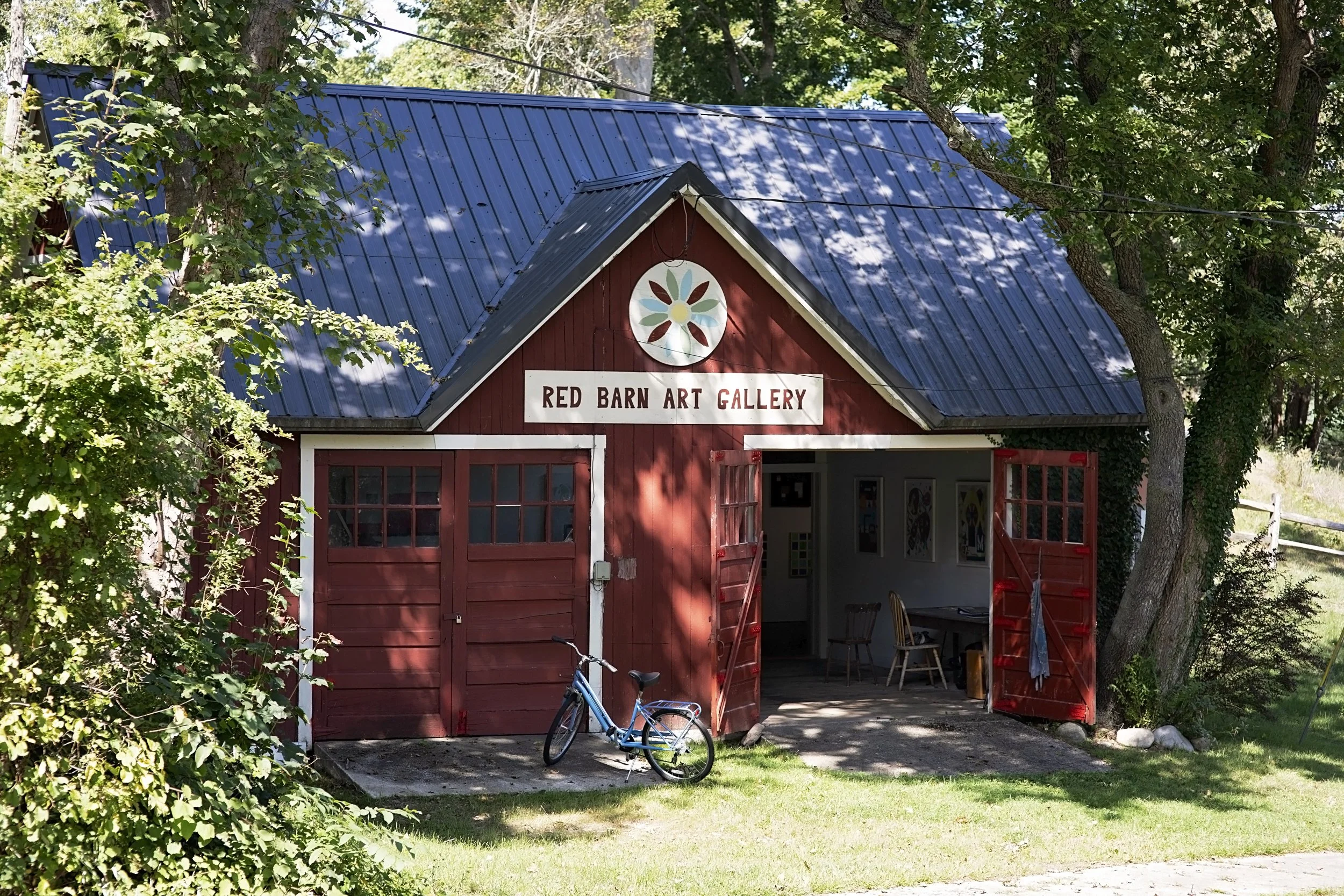 Red barn art gallery with open red doors, bicycle parked outside, surrounded by green trees and grass, blue roof, sign reading 'Red Barn Art Gallery' above entrance.
