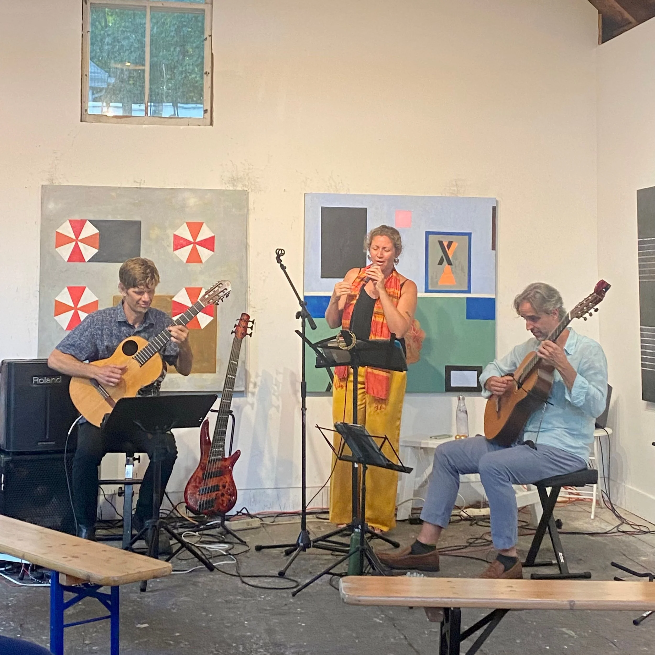 Three musicians performing indoors. Two men are playing acoustic guitars, and a woman is playing a small wind instrument, possibly a flute or recorder. They are standing and sitting near a white wall with abstract paintings behind them.