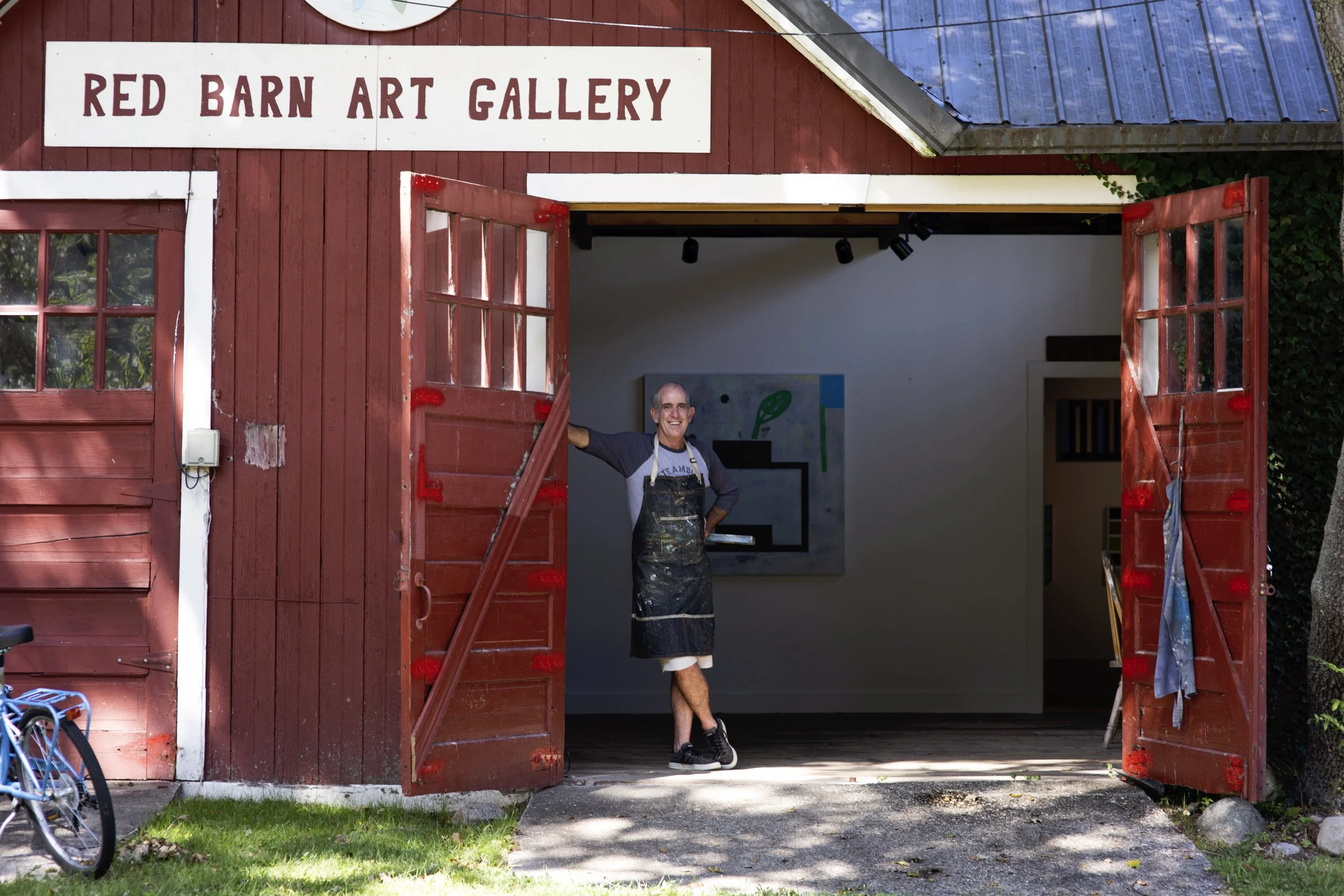 Charles Thomas O'Neil standing inside the Red Barn Art Gallery, smiling, with open red barn doors, artwork on the wall, and a bicycle outside.