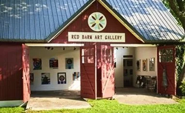 Red barn art gallery with open doors displaying artwork inside, sign above entrance reading "Red Barn Art Gallery," metal roof, trees nearby.