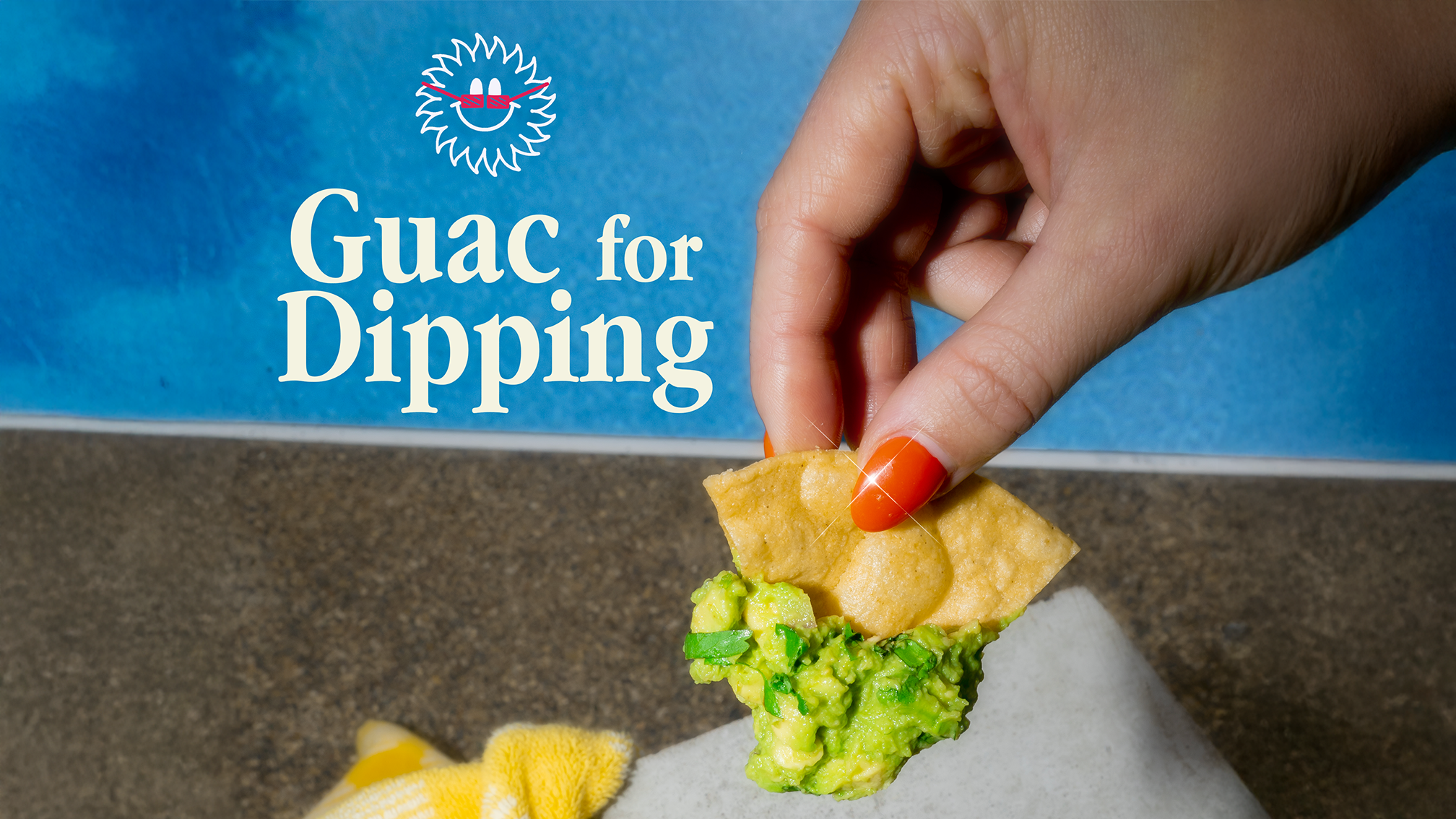 A hand with orange nail polish holding a tortilla chip topped with mashed avocado and guacamole, with a blue sign in the background reading "Guac for Dipping" and a smiley face logo above the text.