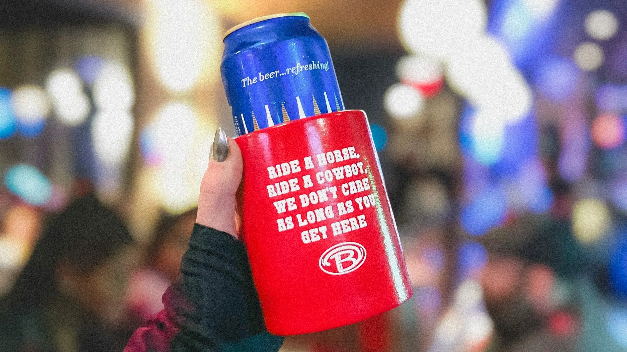Person holding a can of beer in a red koozie that has a humorous phrase and the Budweiser logo, with a blurry background of colorful lights and people.
