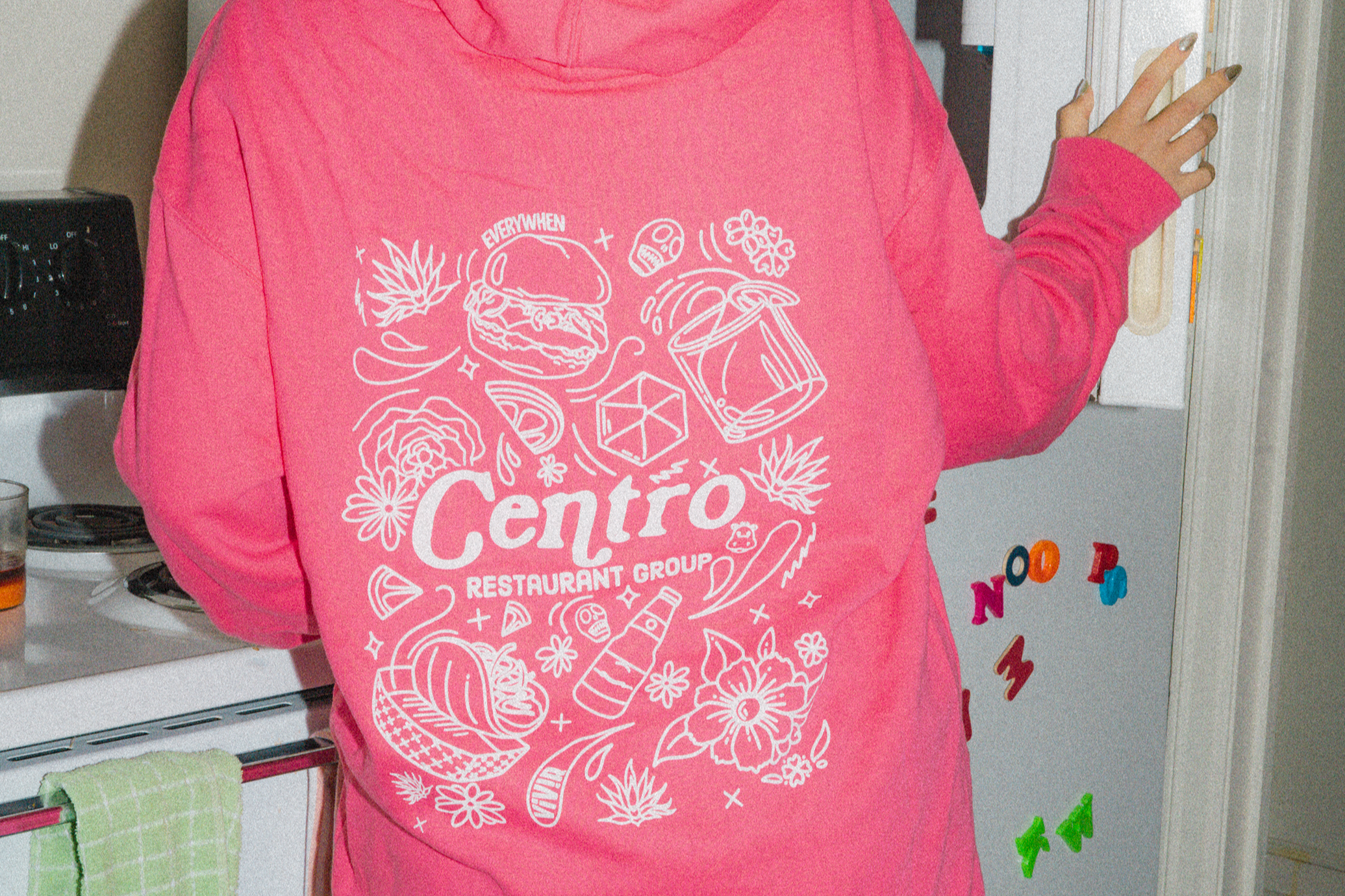 Person wearing a pink sweatshirt with white graphic design and text reading "Centro Restaurant Group," standing in a kitchen near a refrigerator with colorful magnetic letters.