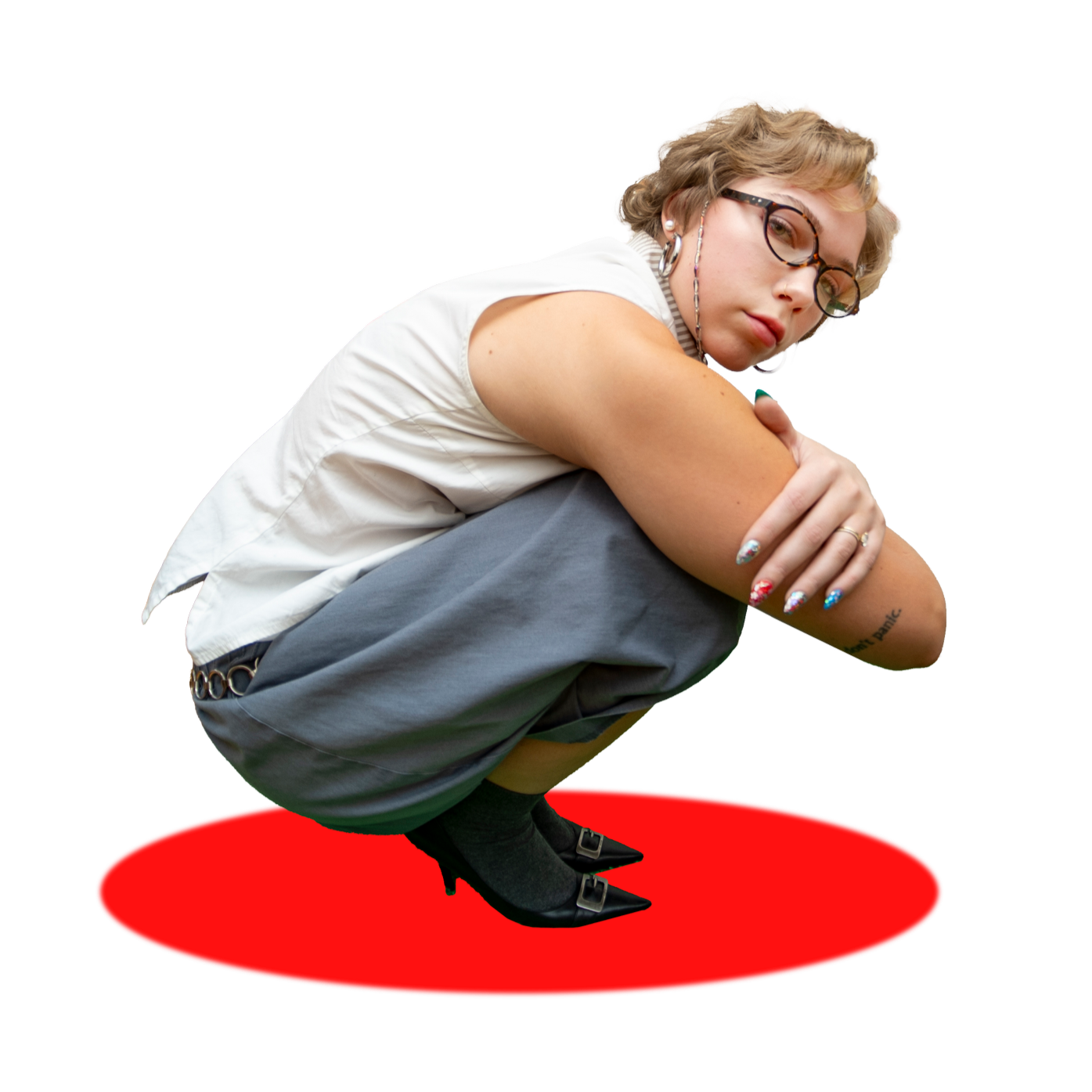 A young woman with blond hair, glasses, and tattoos squats on a red circle, wearing a white sleeveless top, gray pants, and black heels, looking at the camera.