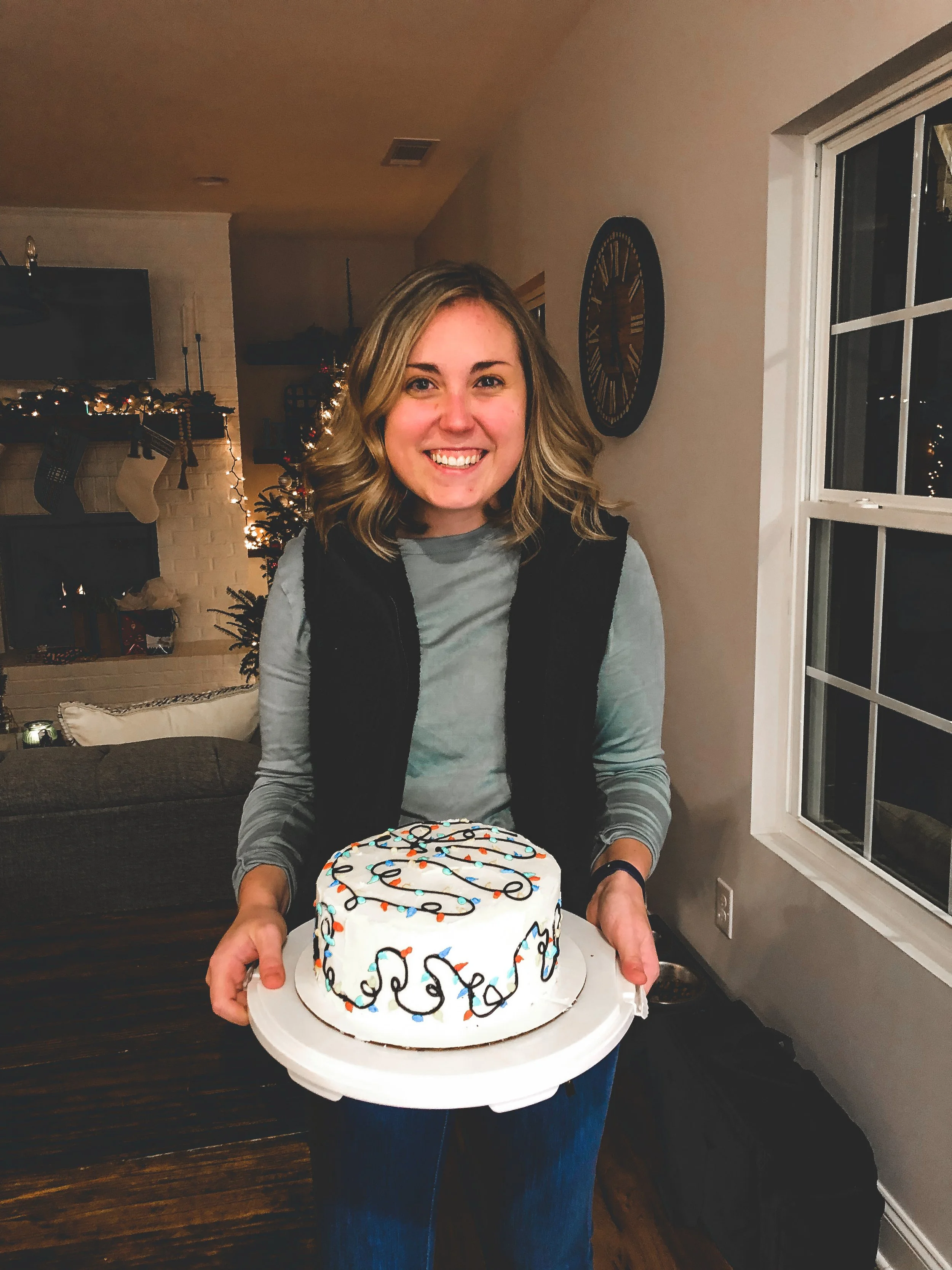 A smiling young woman holding a birthday cake decorated with colorful icing and lights in a cozy living room with a Christmas tree in the background.