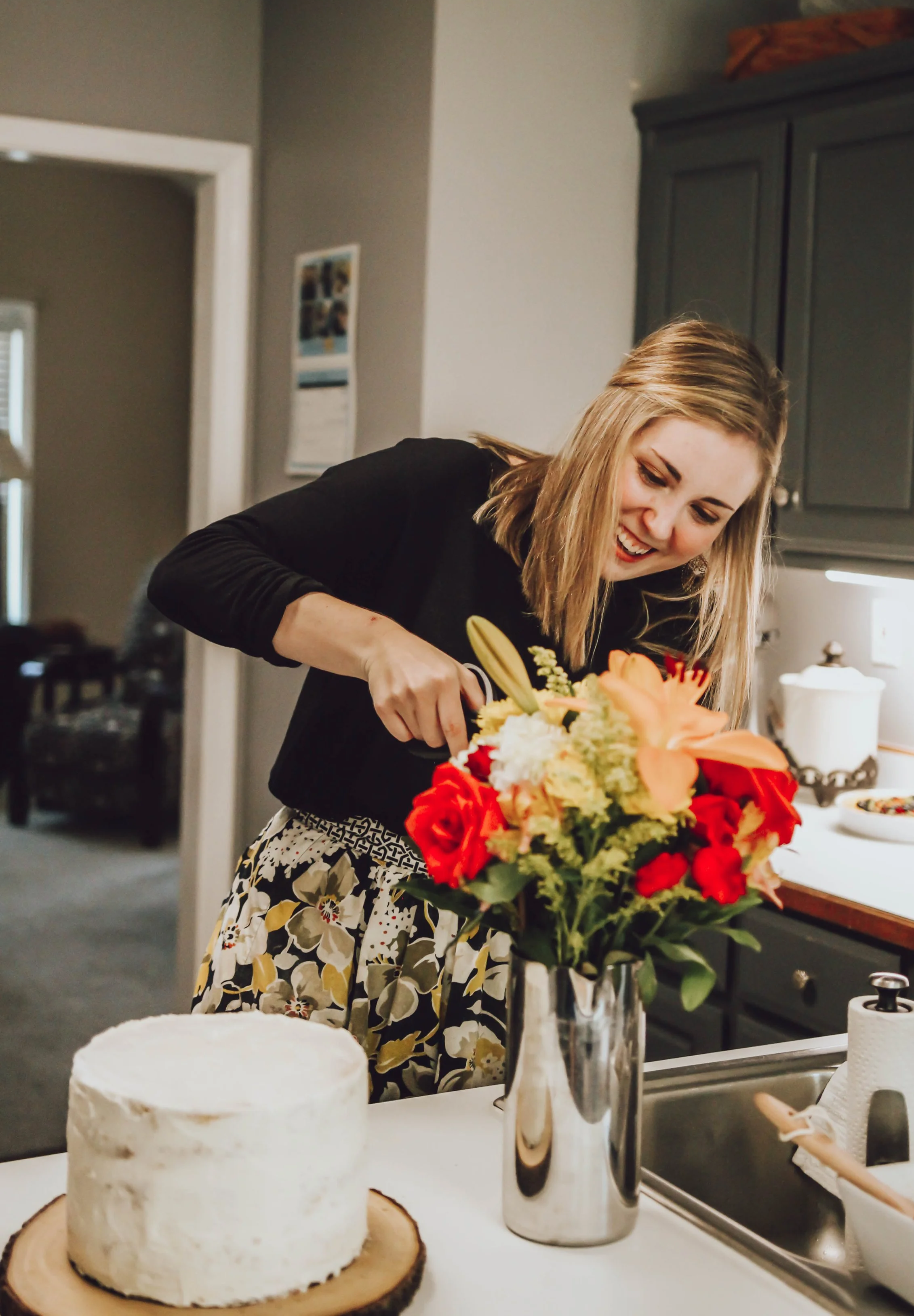 A woman cutting flower bouquet in a kitchen with a white cake, knife, and flowers on the counter.