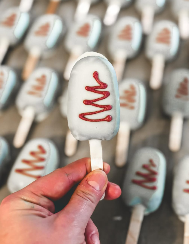 Hand holding a toasted marshmallow ice cream treat with white coating and red drizzle, with more similar treats in the background.