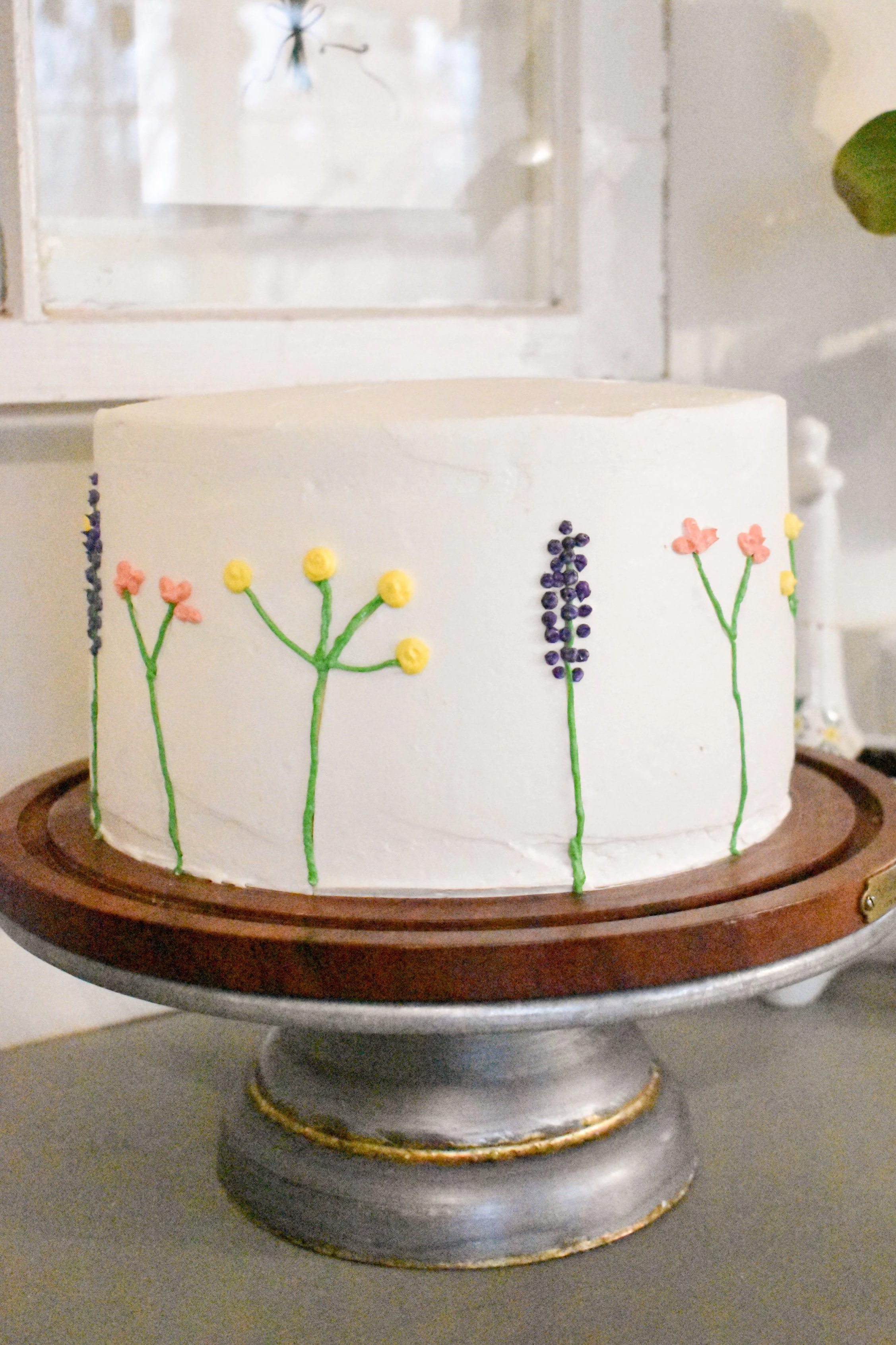A white cake decorated with colorful, simple floral designs, placed on a silver and wooden cake stand.