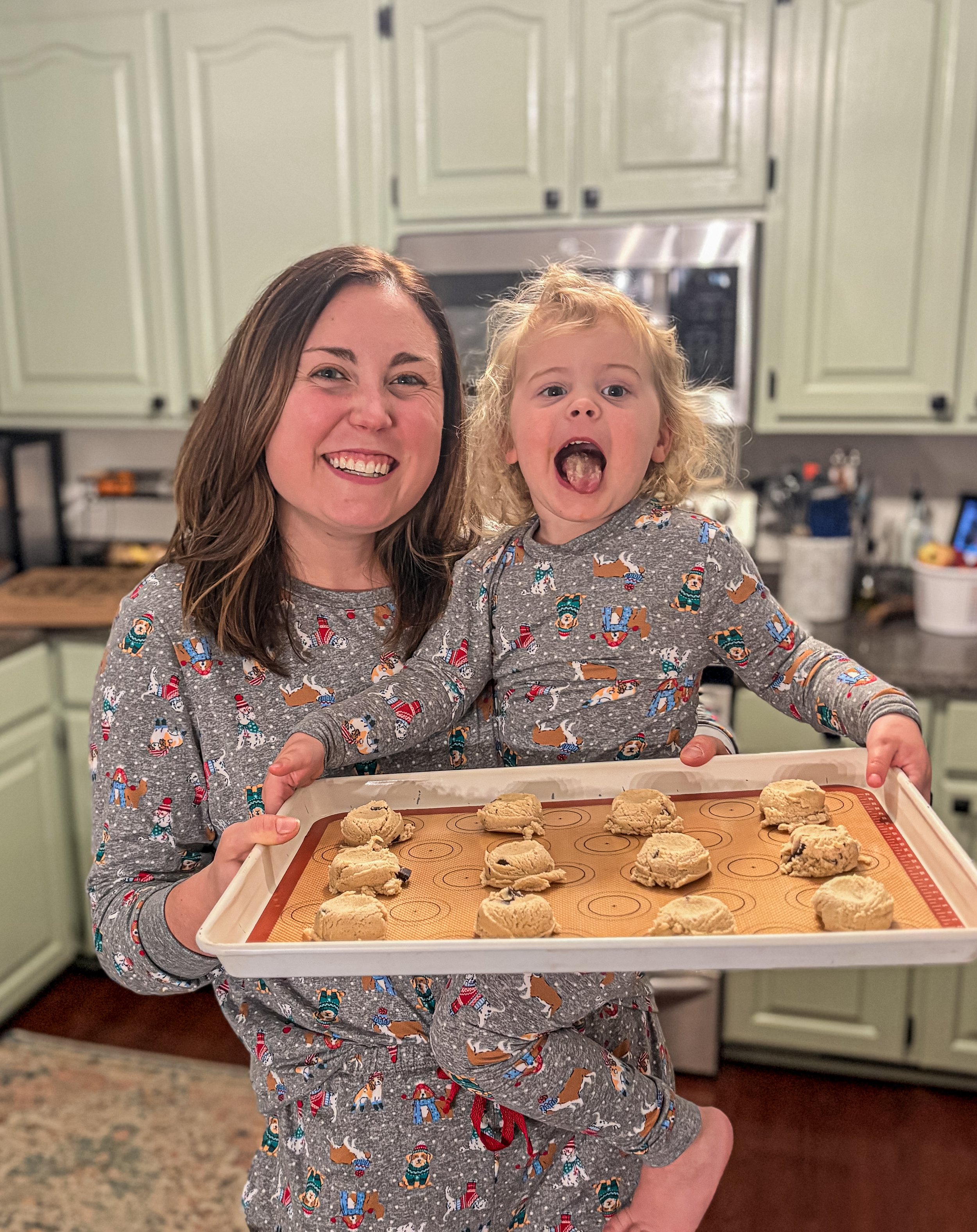 A woman and a young girl in matching holiday pajamas holding a tray of cookie dough balls in a kitchen.