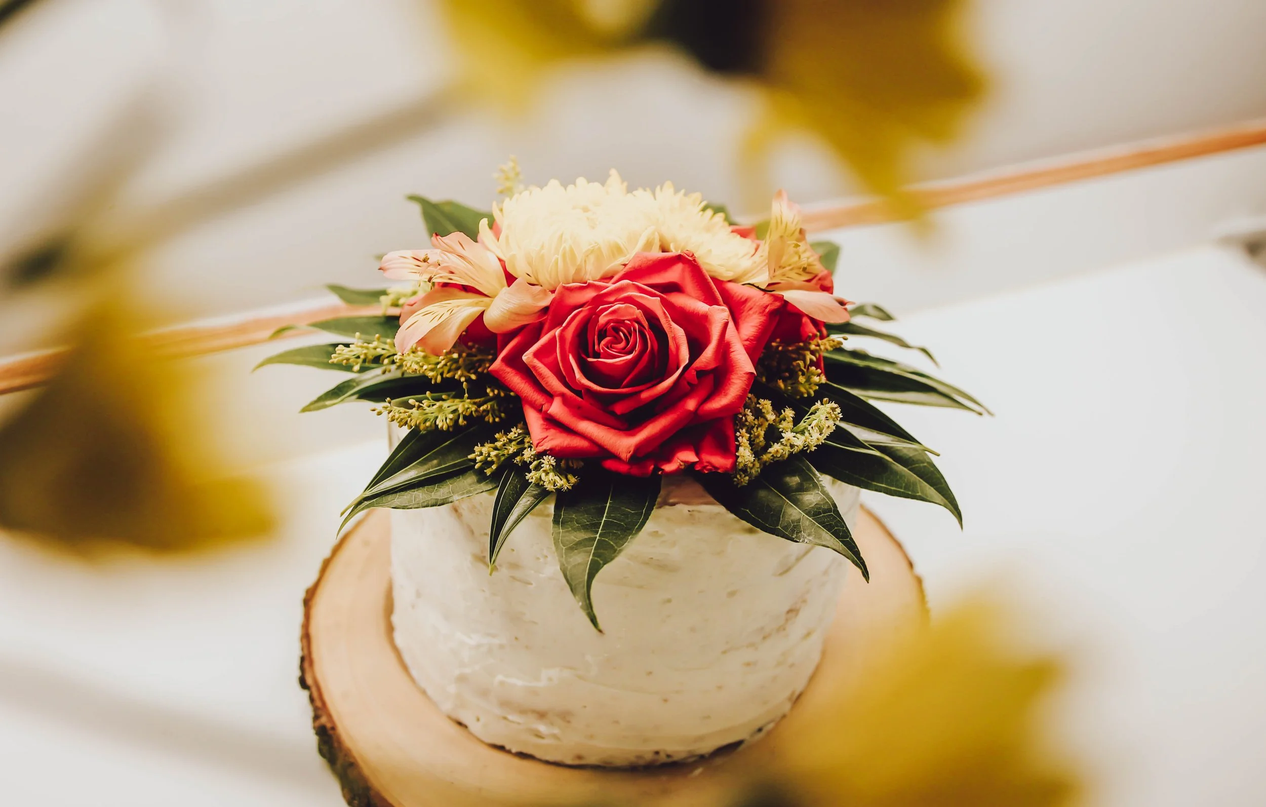 A floral arrangement on top of a white cake, featuring a large red rose, a pink flower, cream-colored blooms, and green leaves, with the cake placed on a round wooden slice.