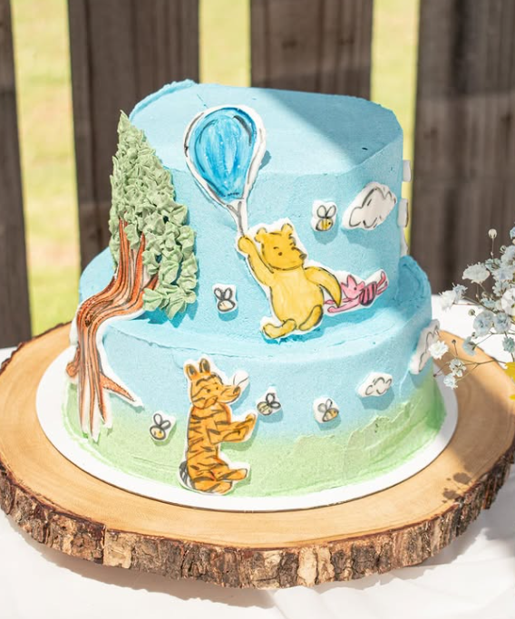 Two-tiered cake with Winnie the Pooh and Tigger decorations, blue sky, white clouds, bees, a tree, and a pink ribbon in the scene.