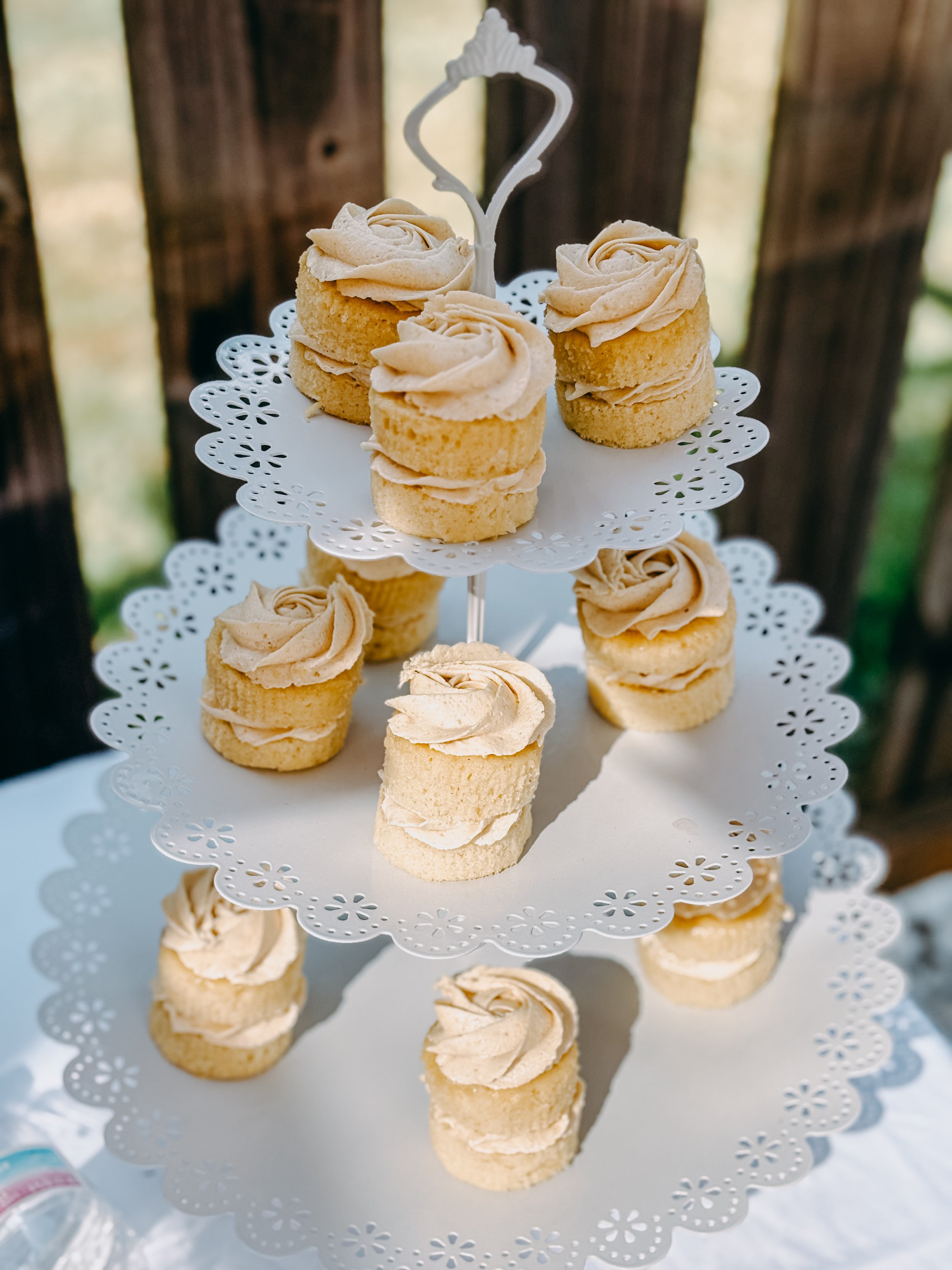 Three-tiered white stand with small vanilla cupcakes topped with swirl of light brown frosting, set outdoors in front of a wooden background.